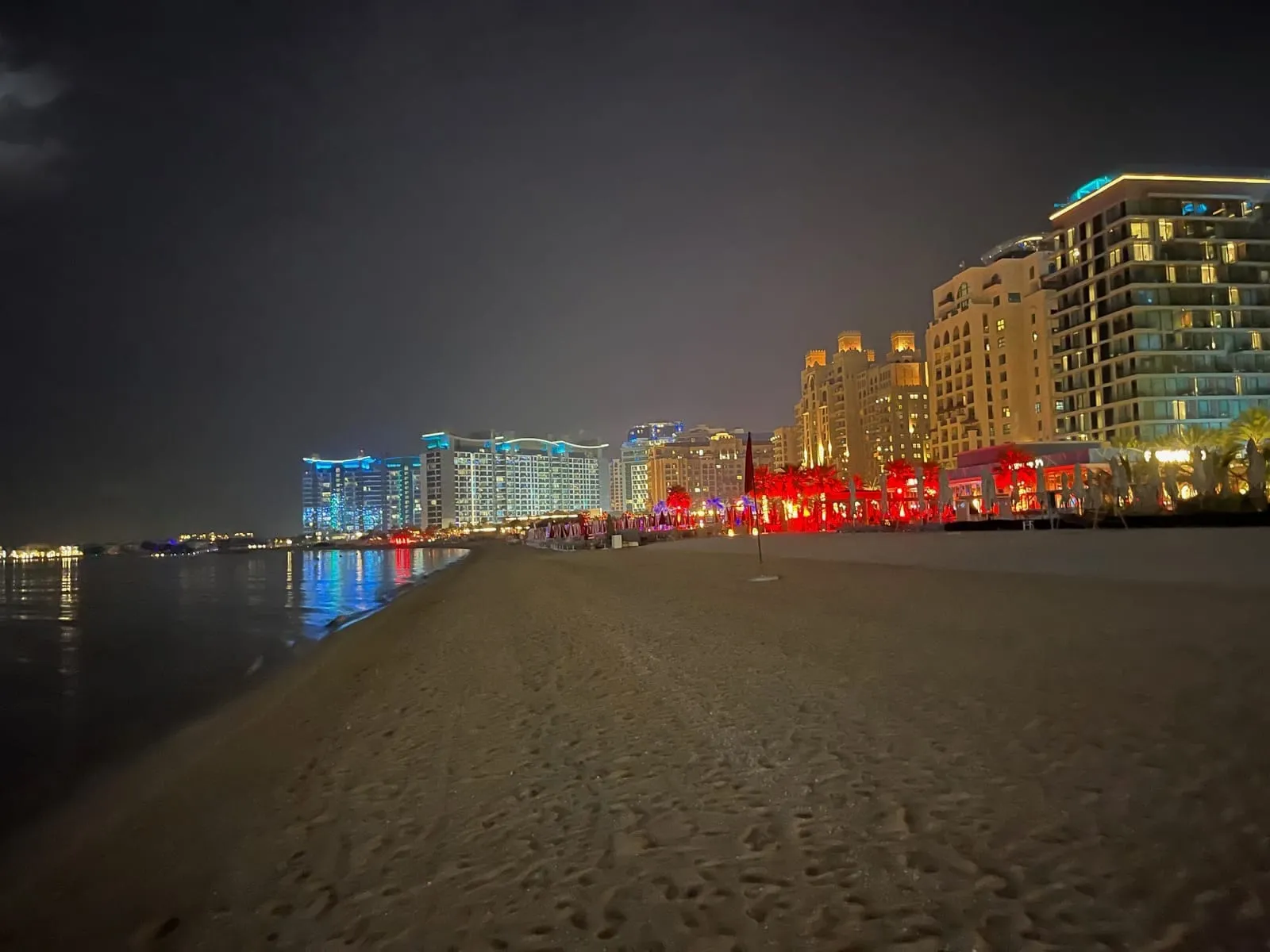 An empty beach at night, footprints in the sand, hotels lit in blue and red along the shore