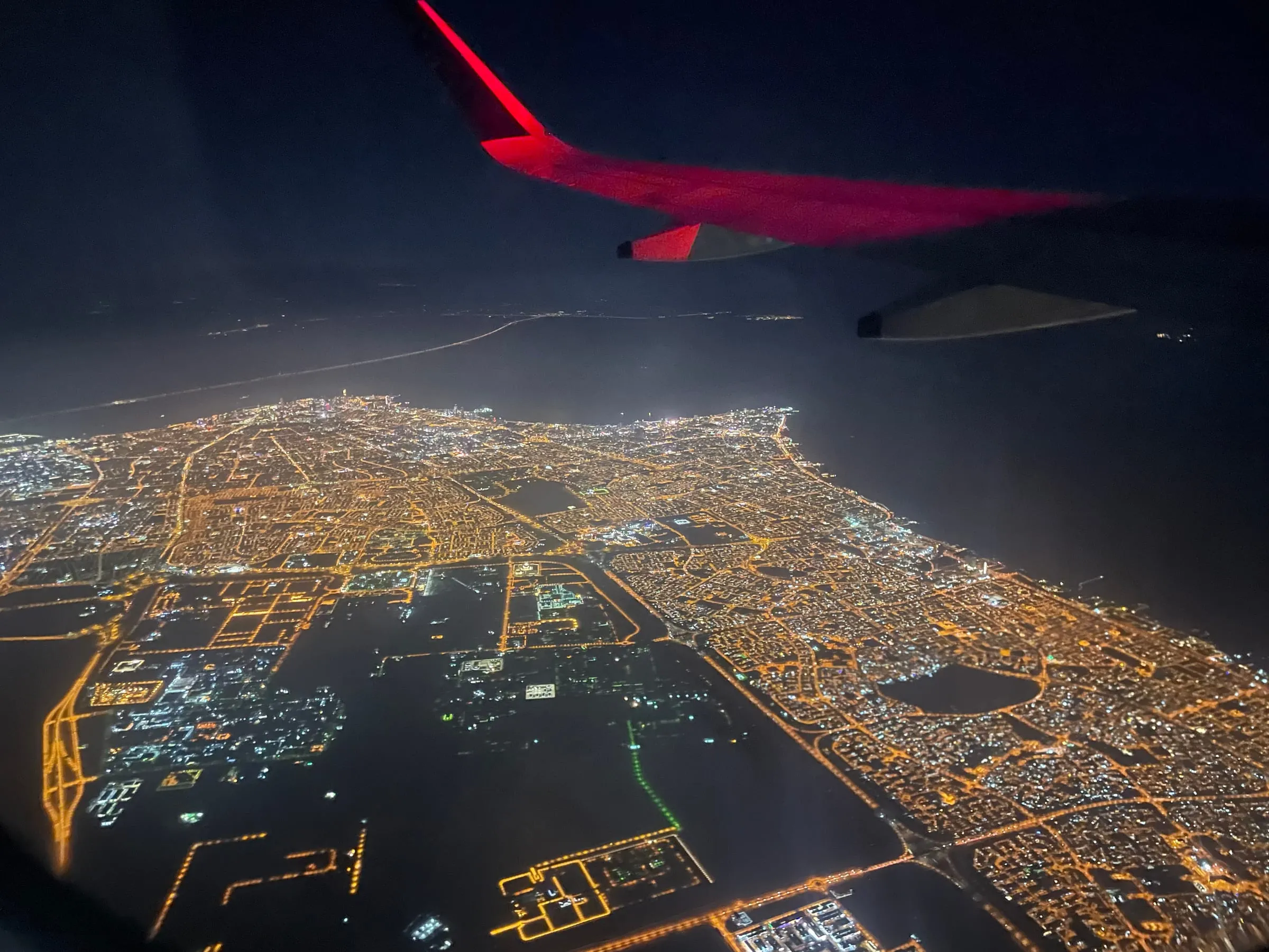 A city's lights at night seen from a plane window, the red wing tip in the corner