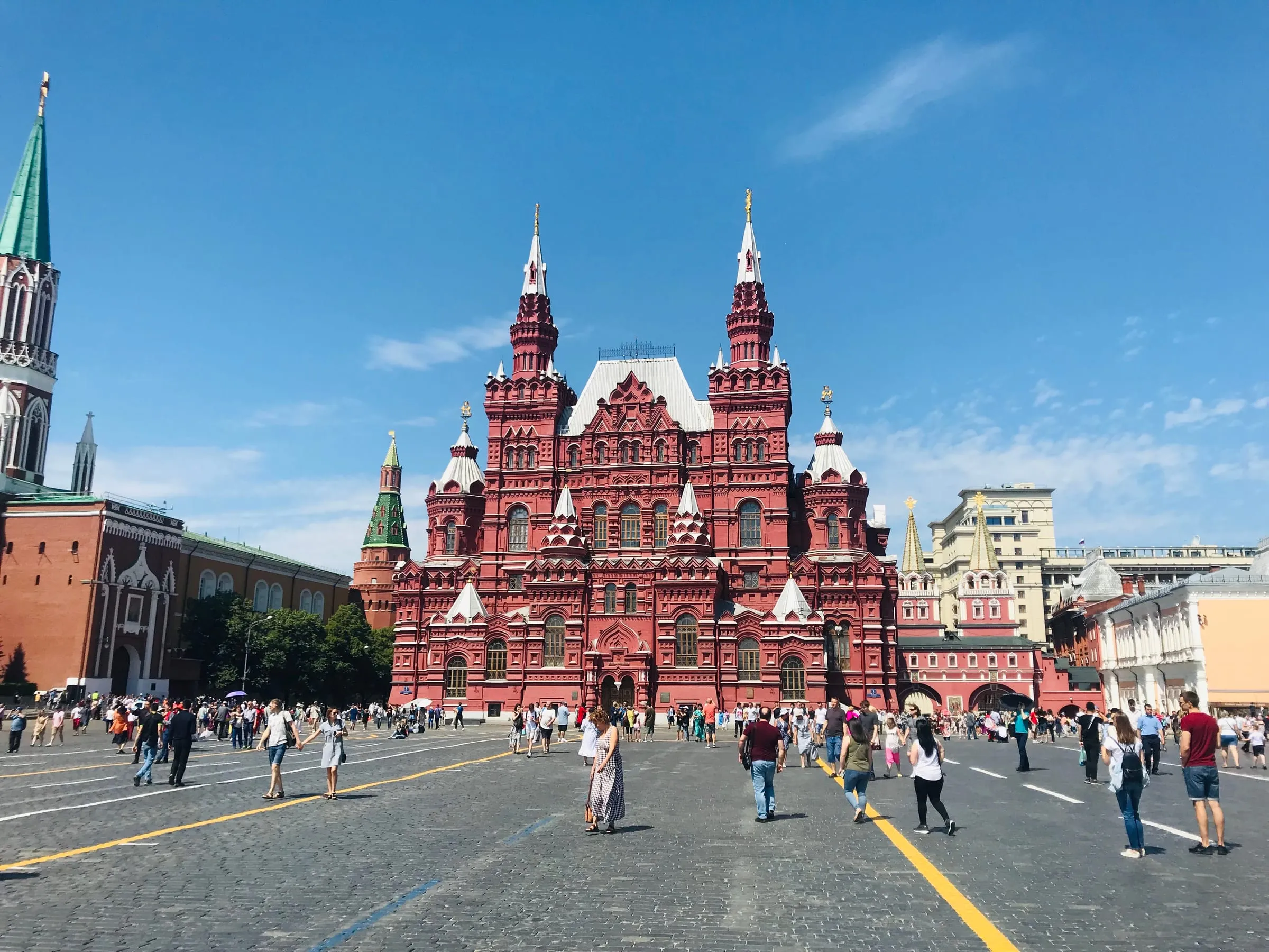 The red brick State Historical Museum on Red Square under a bright blue sky, crowds walking across the square