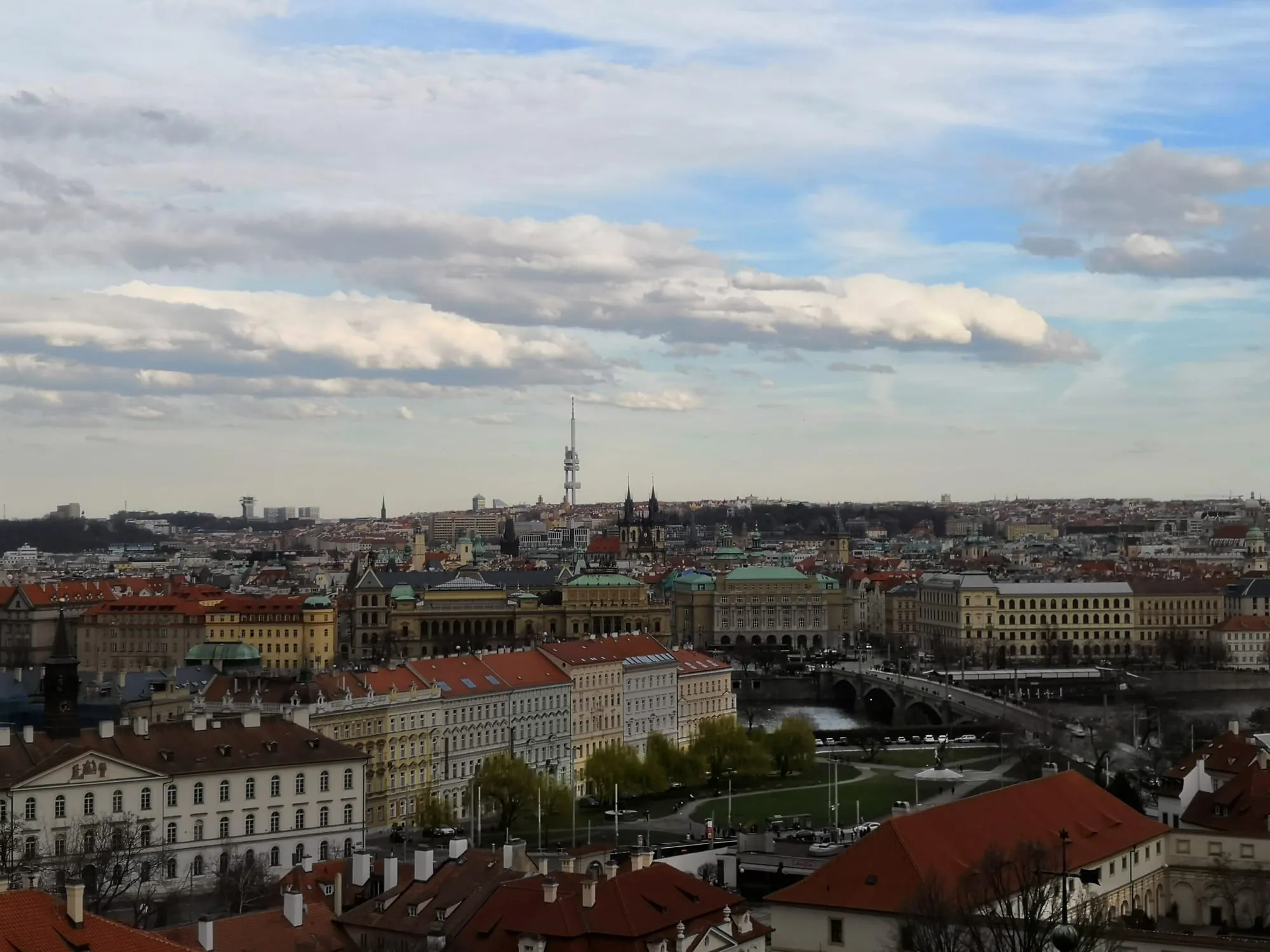 A view over the red rooftops of Prague towards a tall TV tower, under a cloudy sky