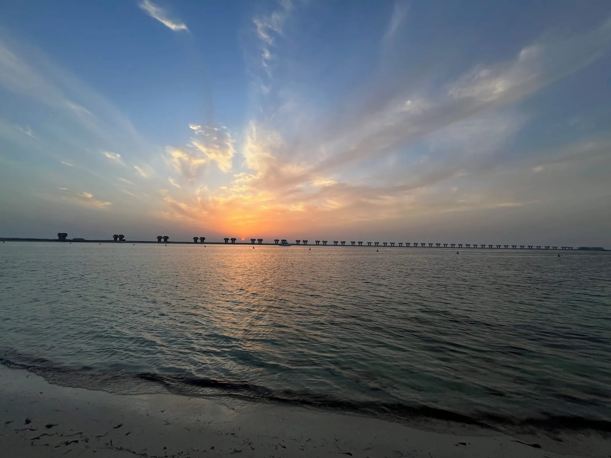 Sunset over calm water from a sandy beach, with a long line of bridge pillars across the horizon