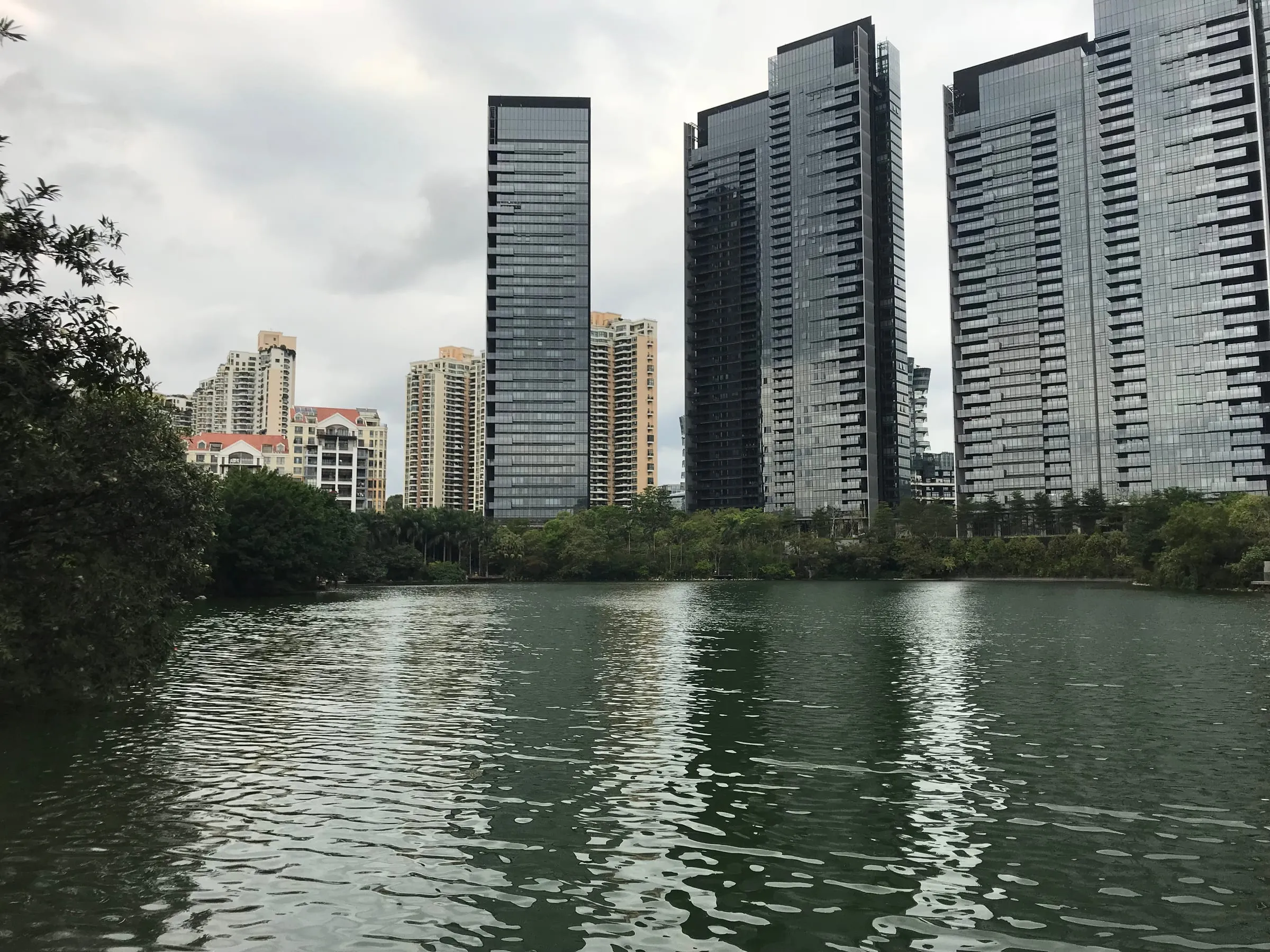 Glass towers rising over a calm lake, trees along the shore under a grey sky