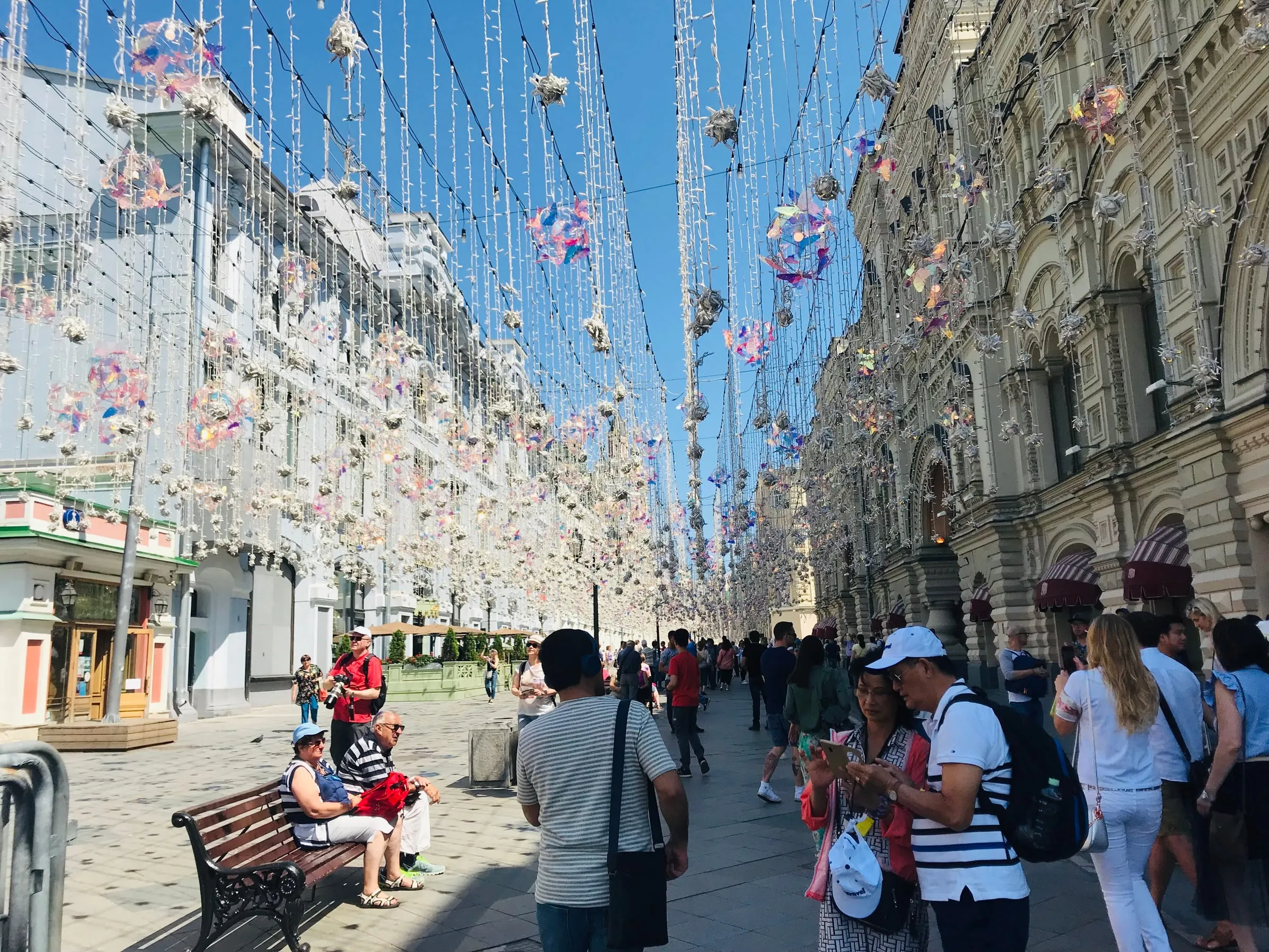 A busy pedestrian street hung with decorations overhead, people walking and sitting on benches