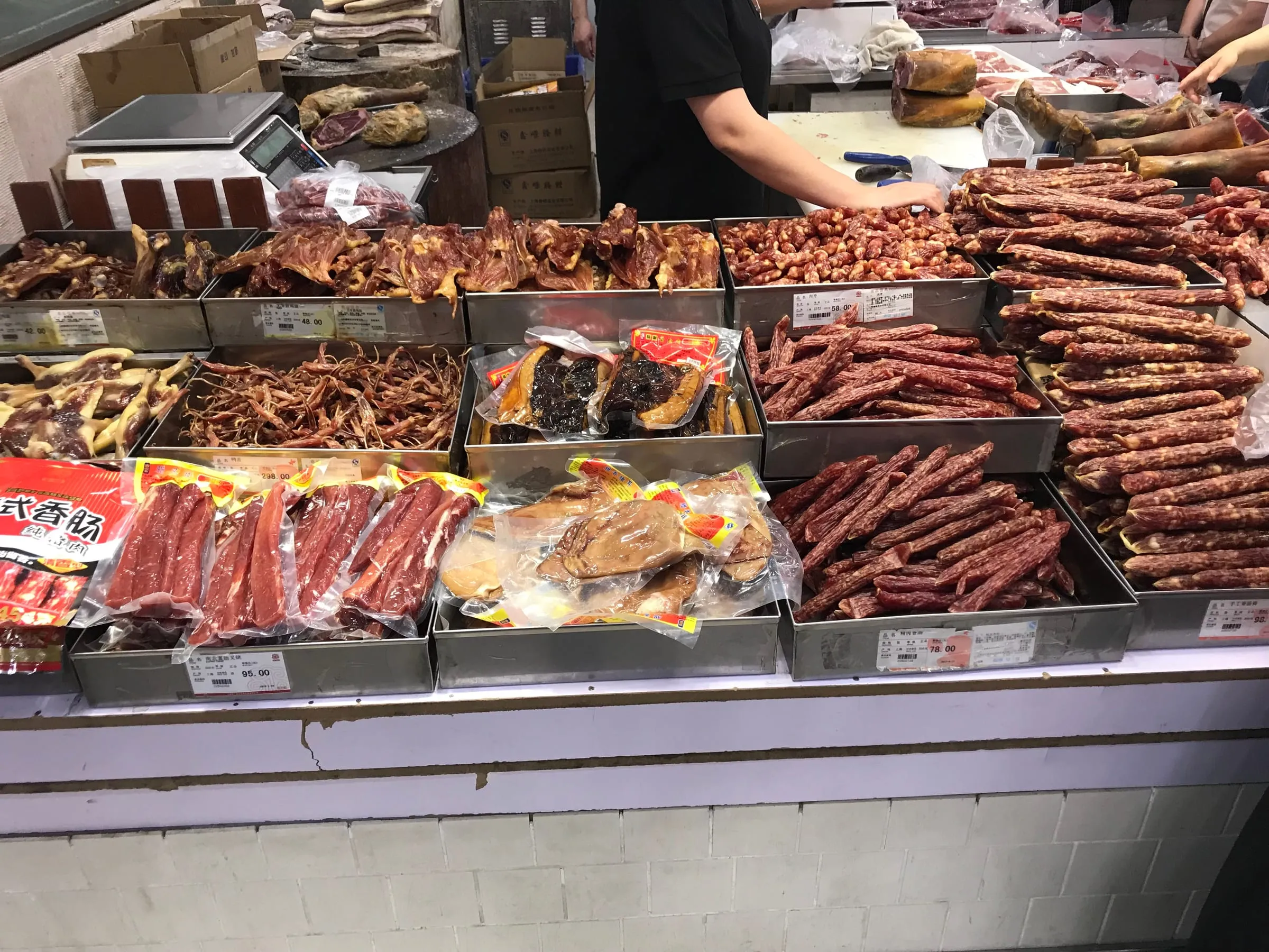 A market counter of dried sausages and cured meats in trays, a scale at the back