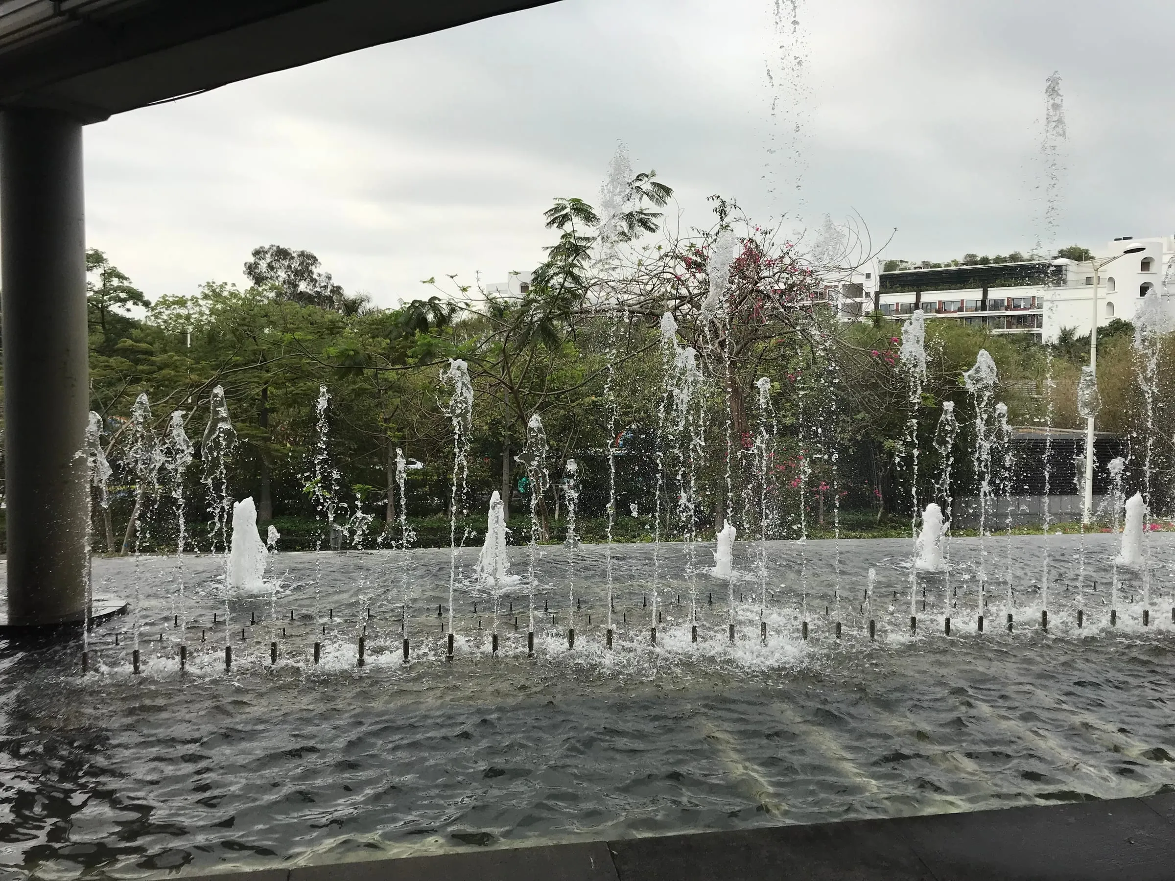 Rows of small fountains in a paved plaza, seen from under a concrete overpass, trees and a white building behind
