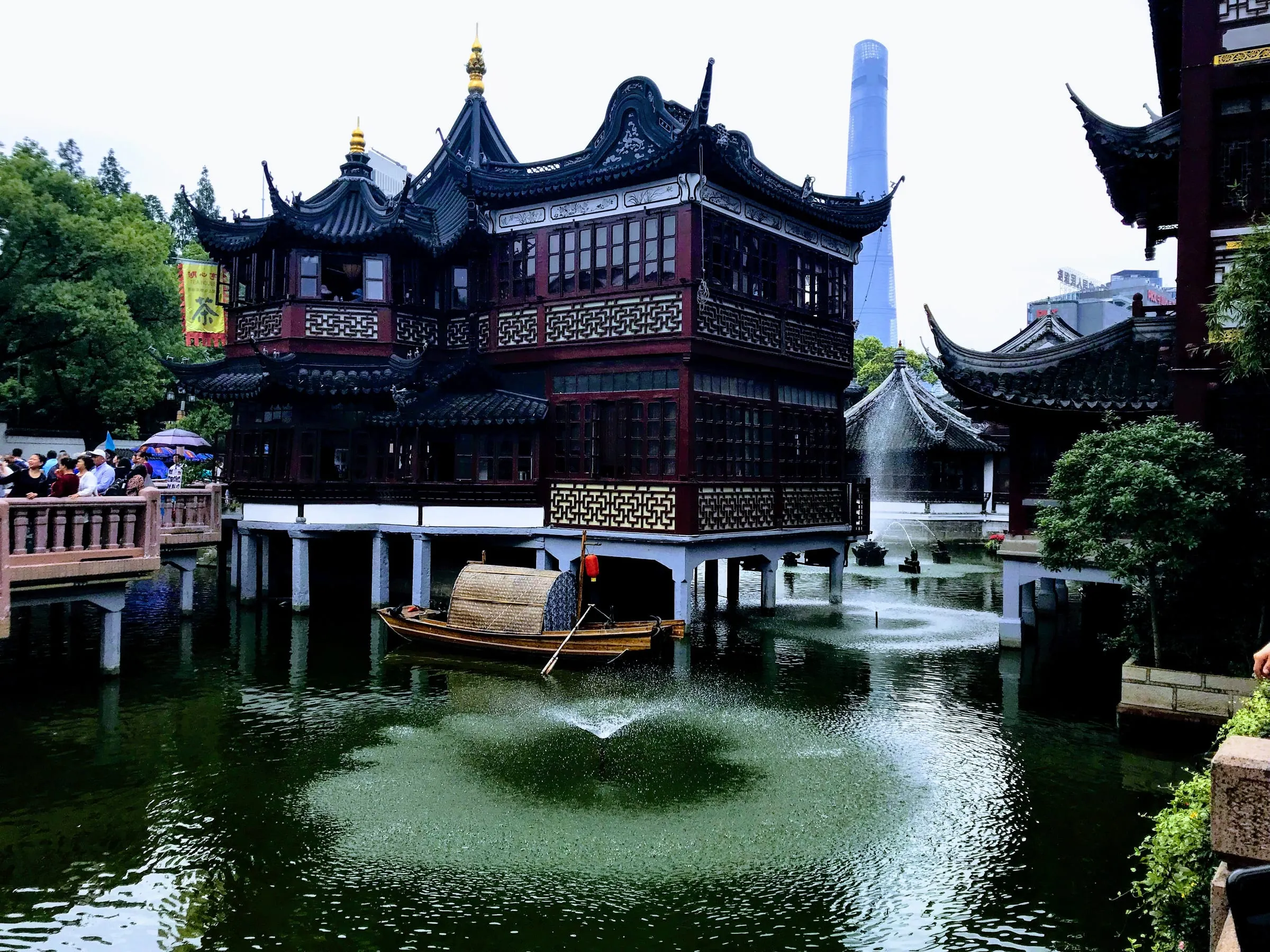 A traditional pavilion with upturned roofs standing over a green pond, a modern tower rising behind