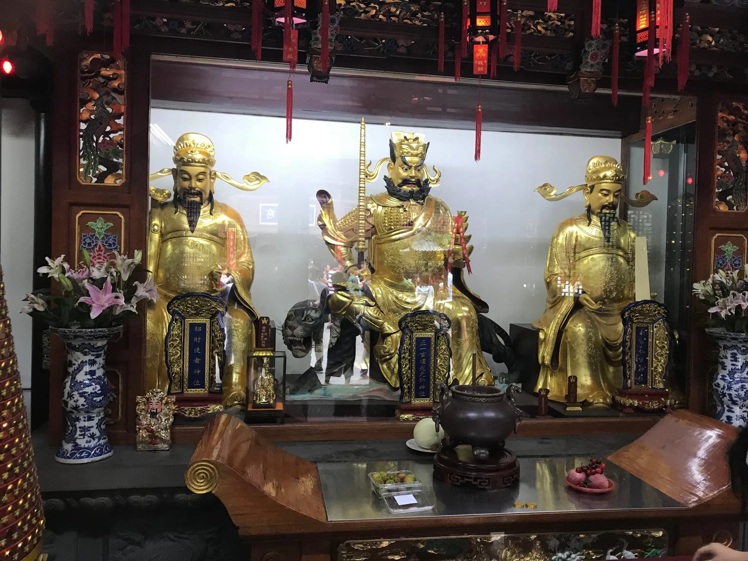 Three gilded seated statues behind an altar with vases of flowers and offerings