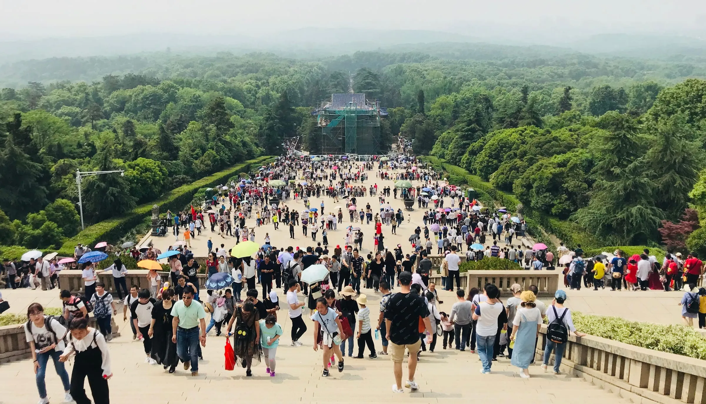 Huge crowds on a wide stone avenue leading through forest towards a distant monument, under a hazy sky