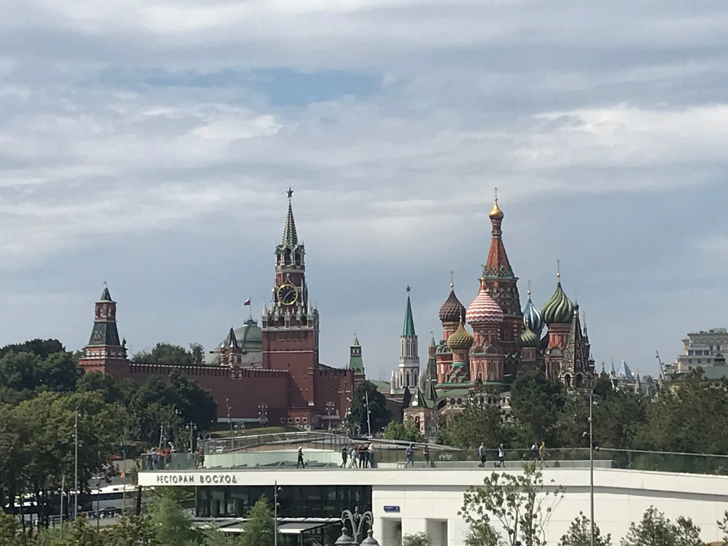 The Kremlin's red towers and St Basil's domes seen from a park across a road, under grey clouds