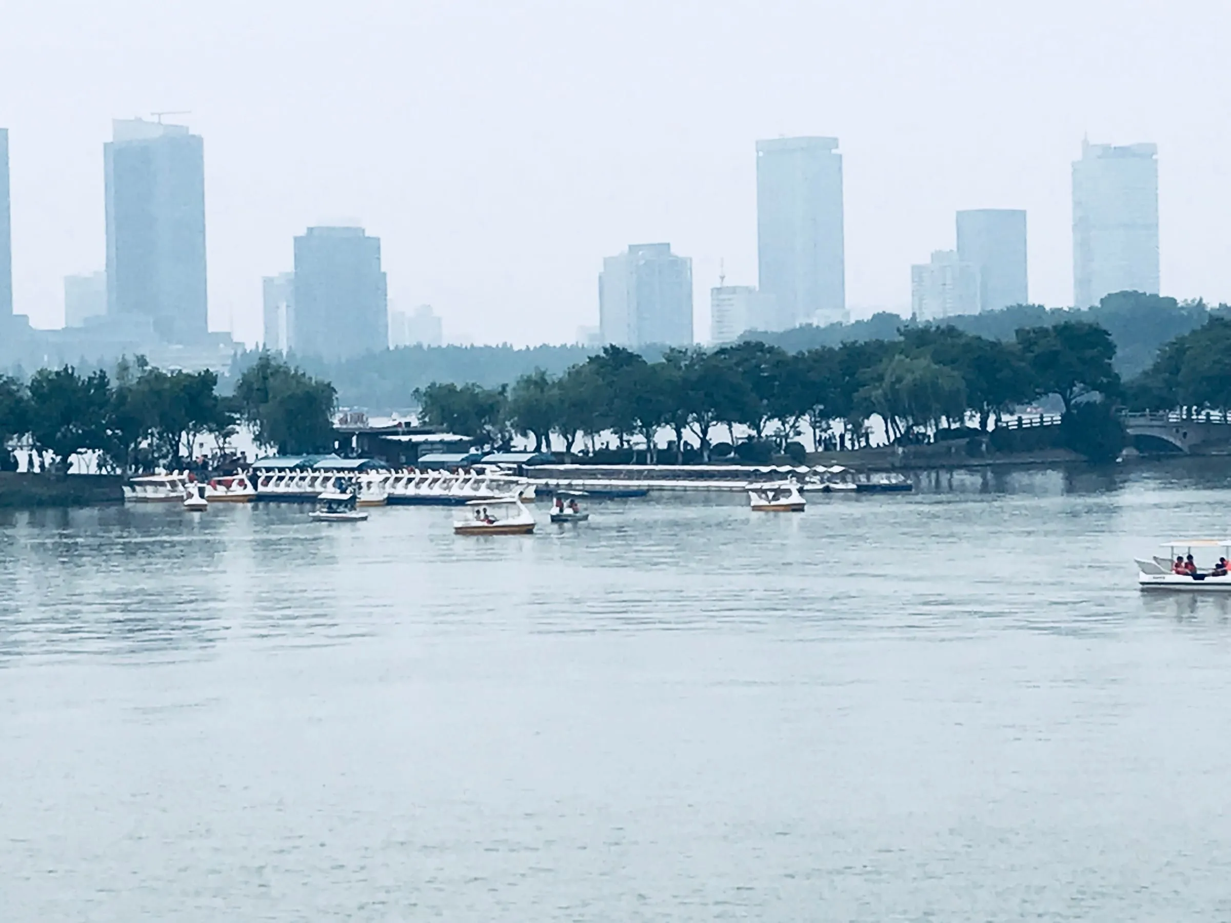 A wide lake with small boats, a line of trees on the far shore and grey towers behind