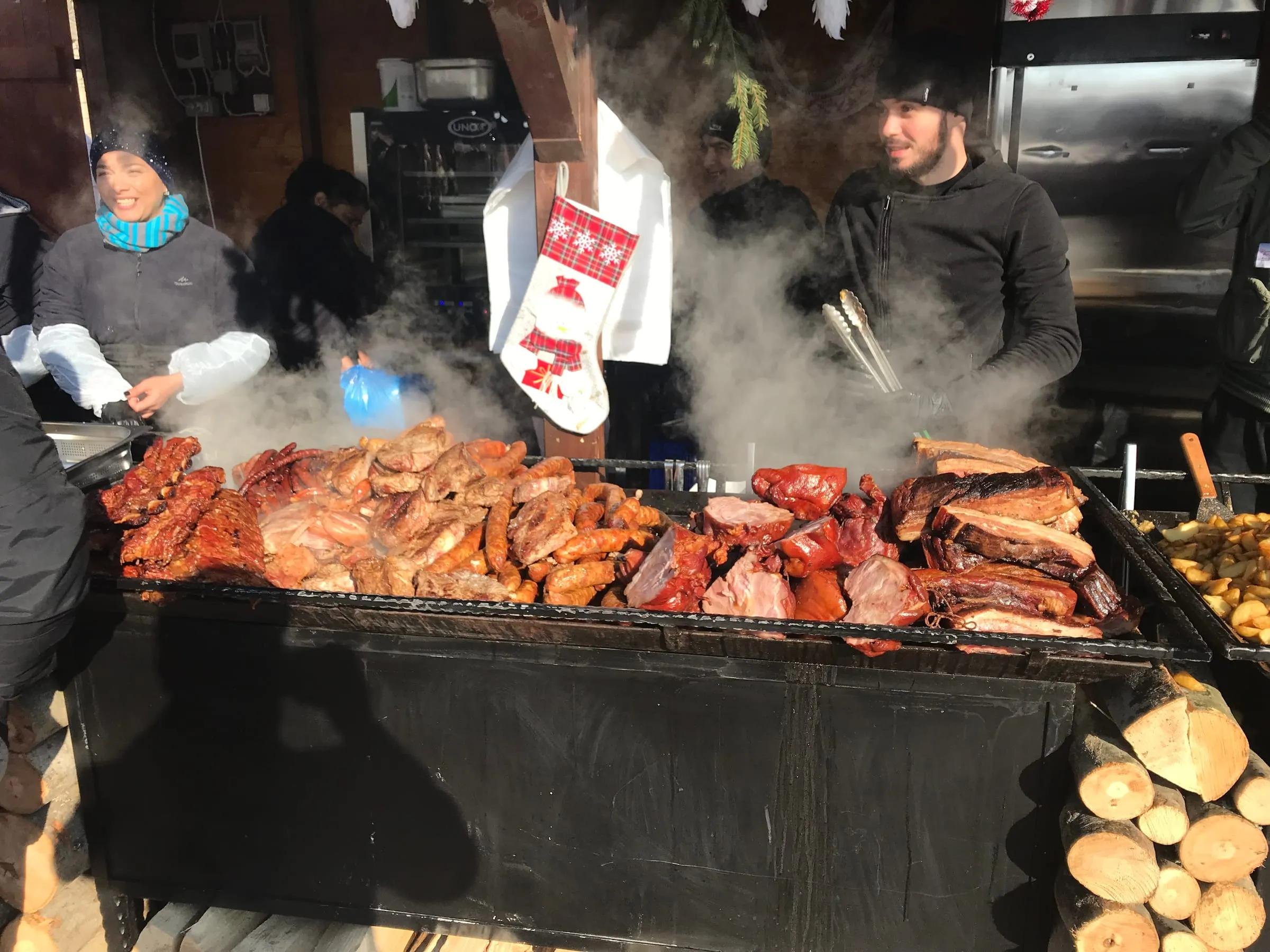 Meat and sausages on a large open grill at a winter market stall, steam rising, a Christmas stocking hanging above