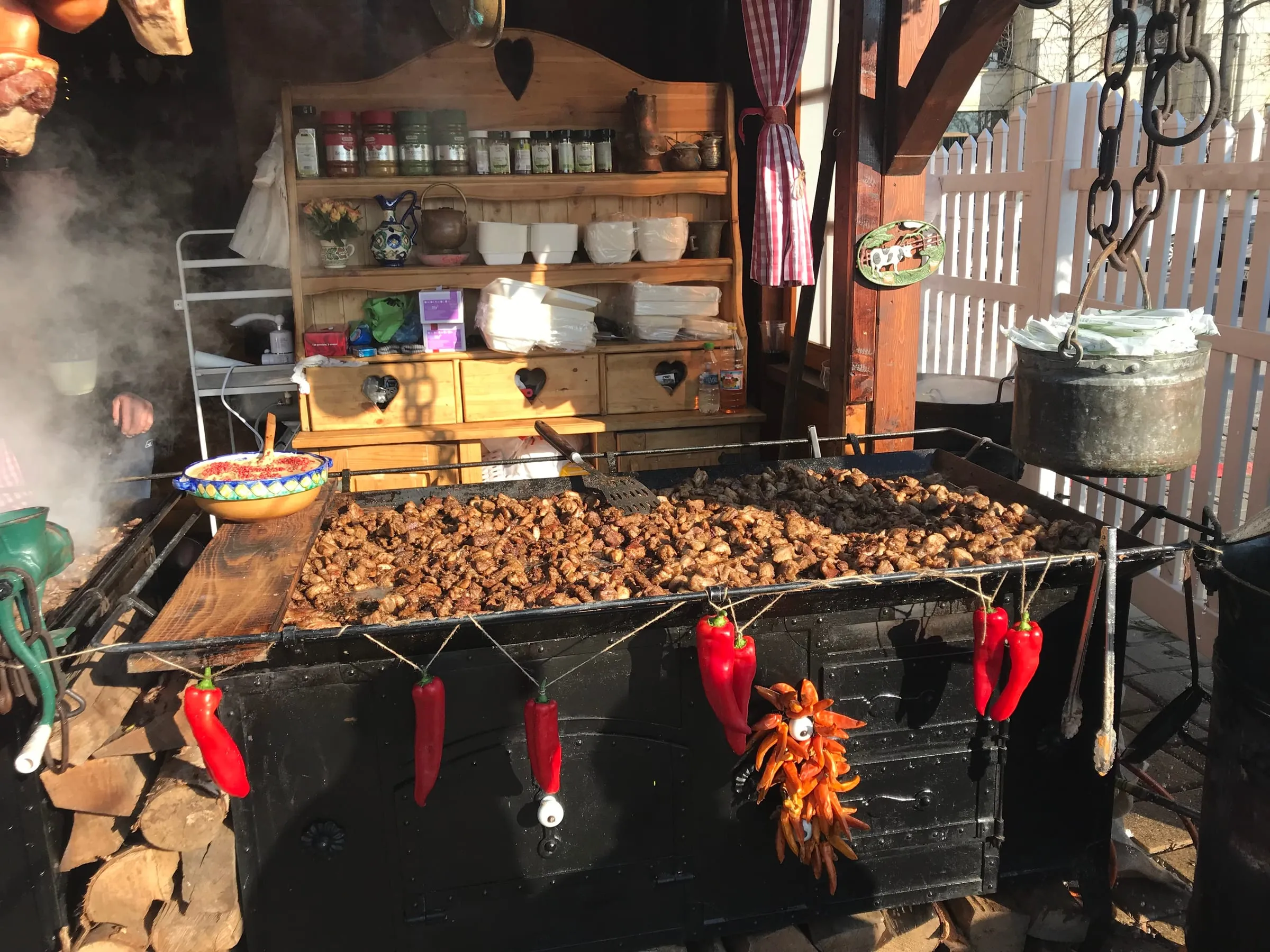 A wide flat pan of cooked food at a wooden market stall, strings of red chillies and firewood stacked below