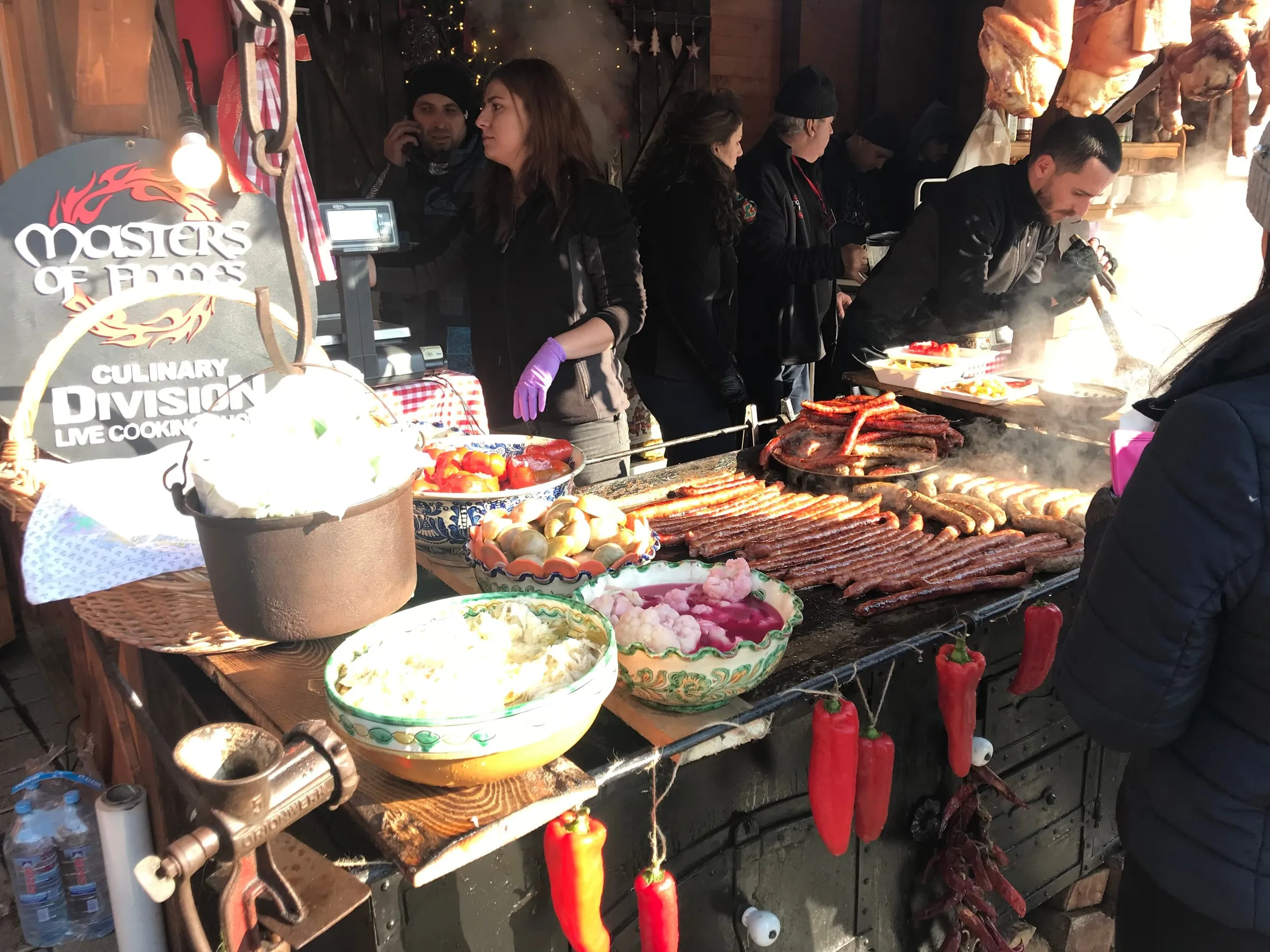 A busy market food stall with bowls of salad, sausages on the grill and red peppers hanging along the front