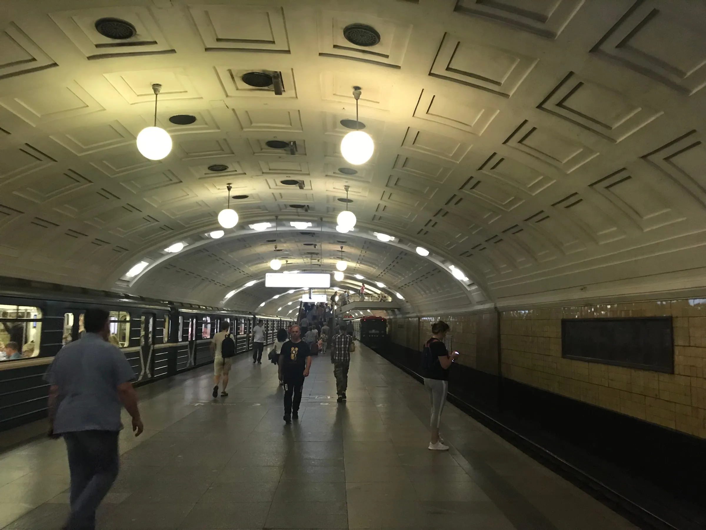 A metro station platform with a curved, ornate ceiling and round lamps, people walking past a train