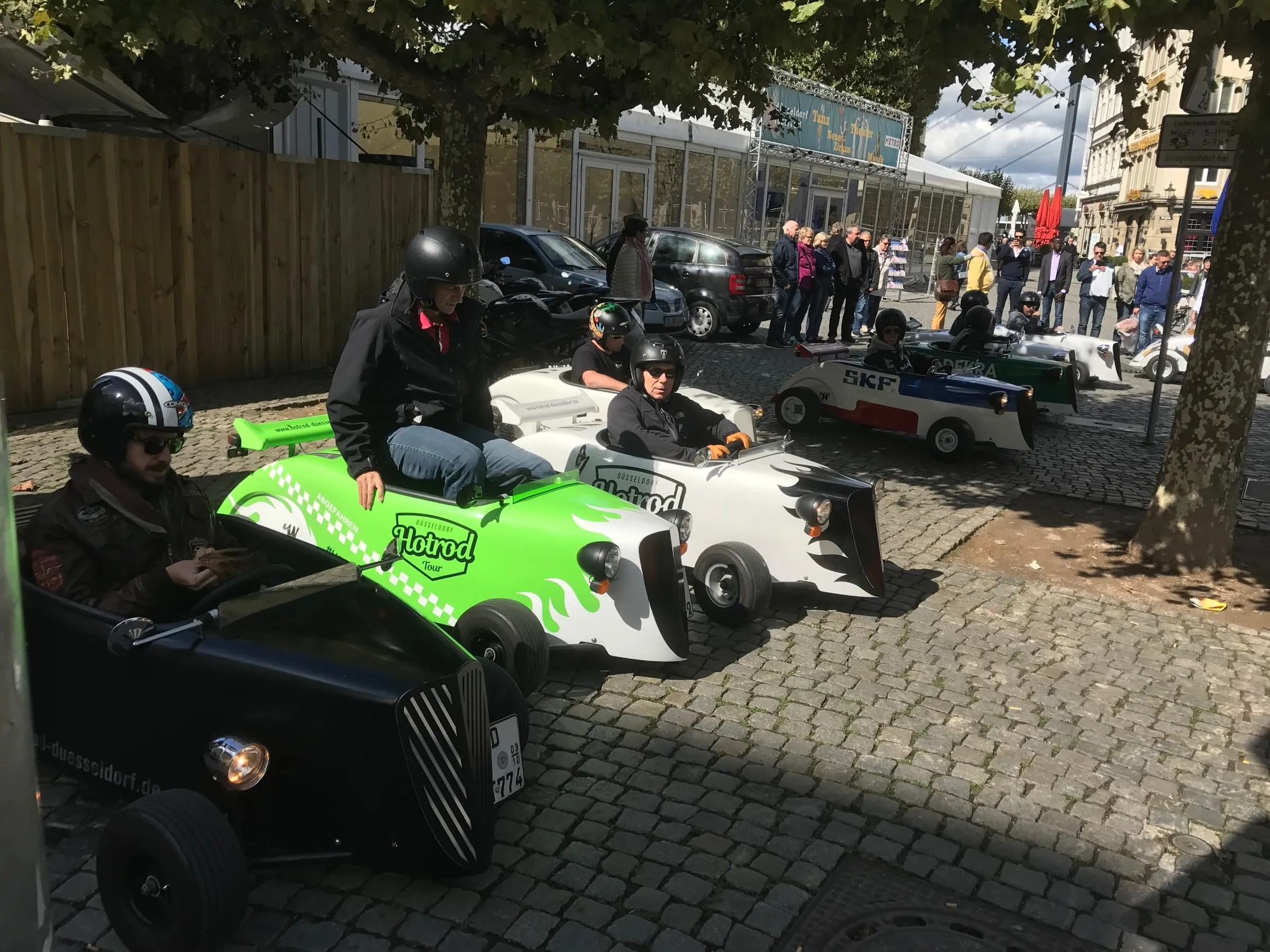People in helmets driving a line of small hot rod cars along a cobbled street