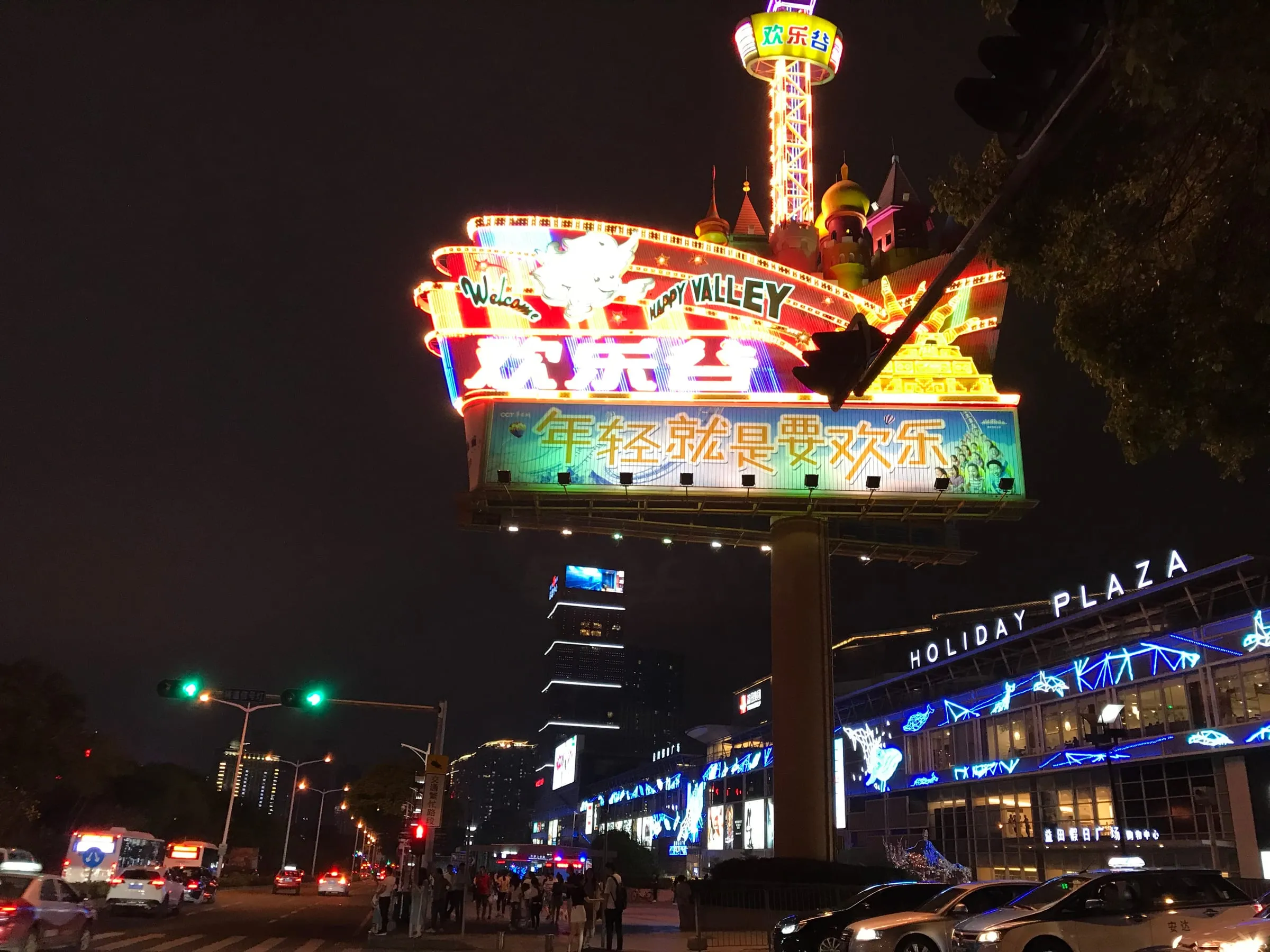 A large neon amusement park sign at night above a busy road, a shopping centre lit in blue behind
