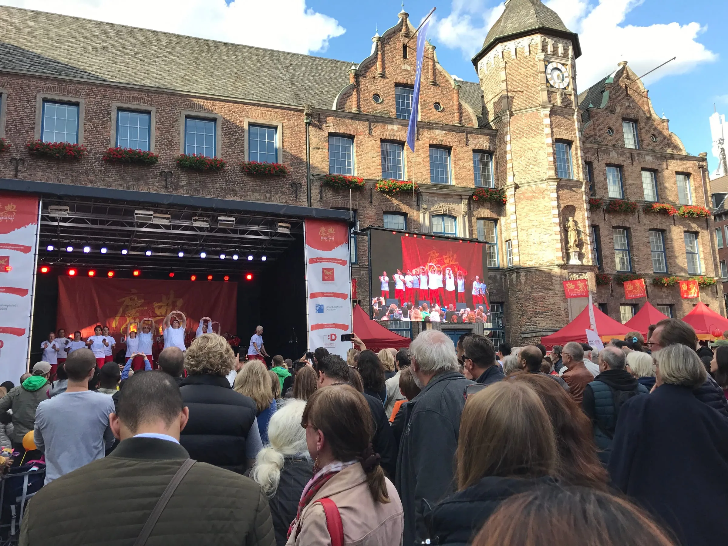 A crowd in front of an outdoor stage in an old town square, a historic brick building with a clock tower behind