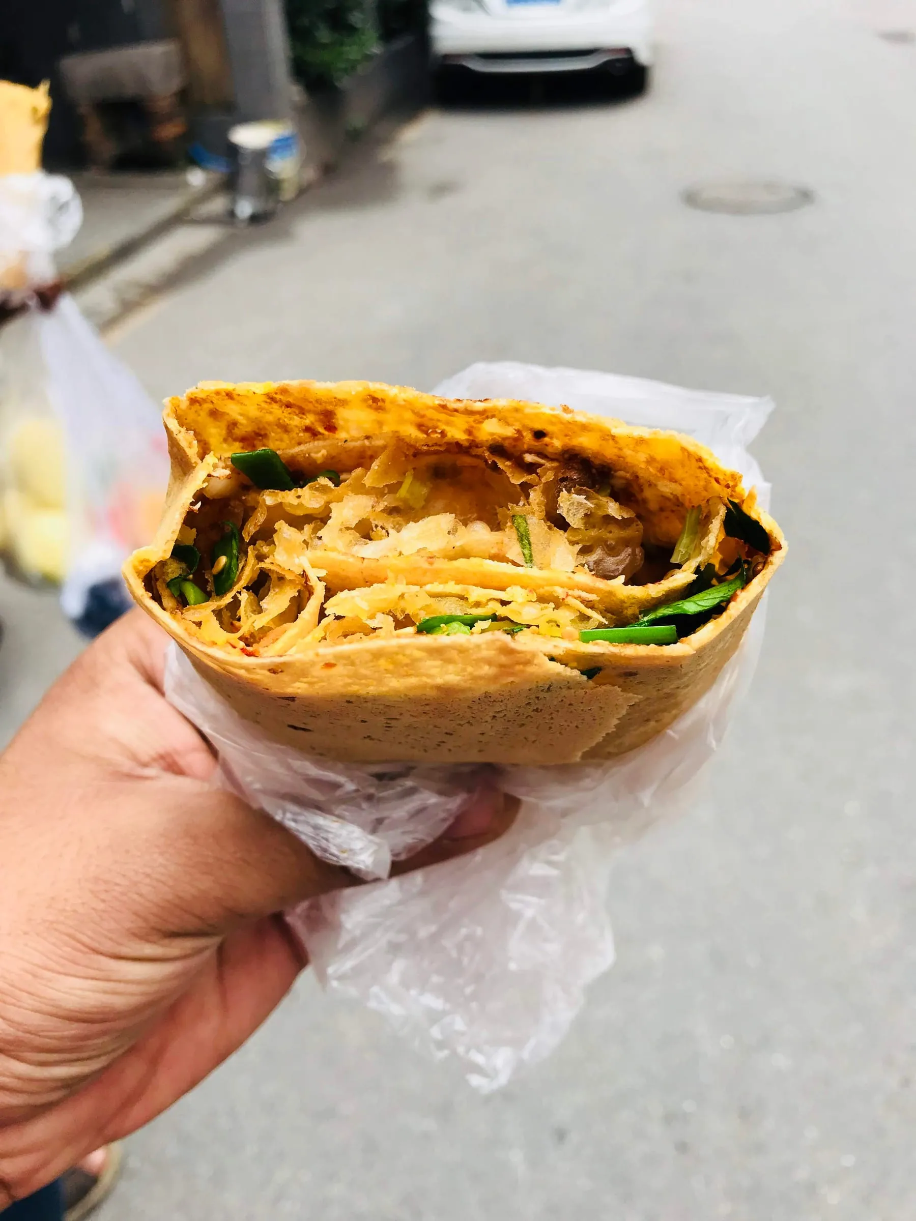 A hand holding a folded street pancake stuffed with crisp filling, the pavement below