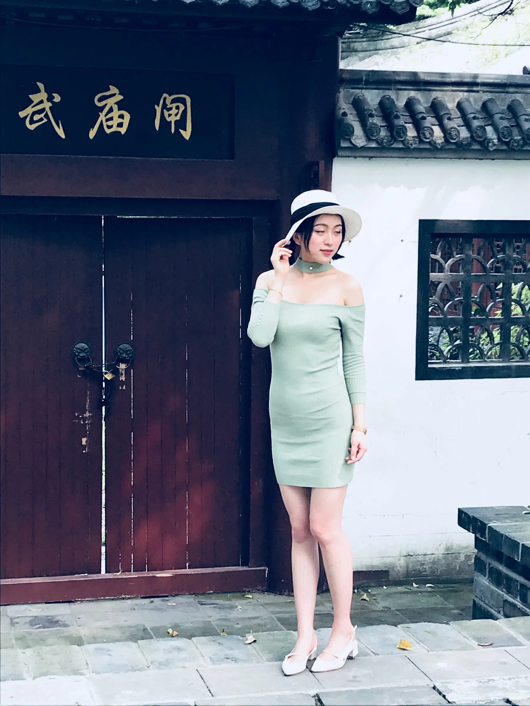 A woman in a pale green dress and white hat posing beside a dark red temple door with a carved sign above