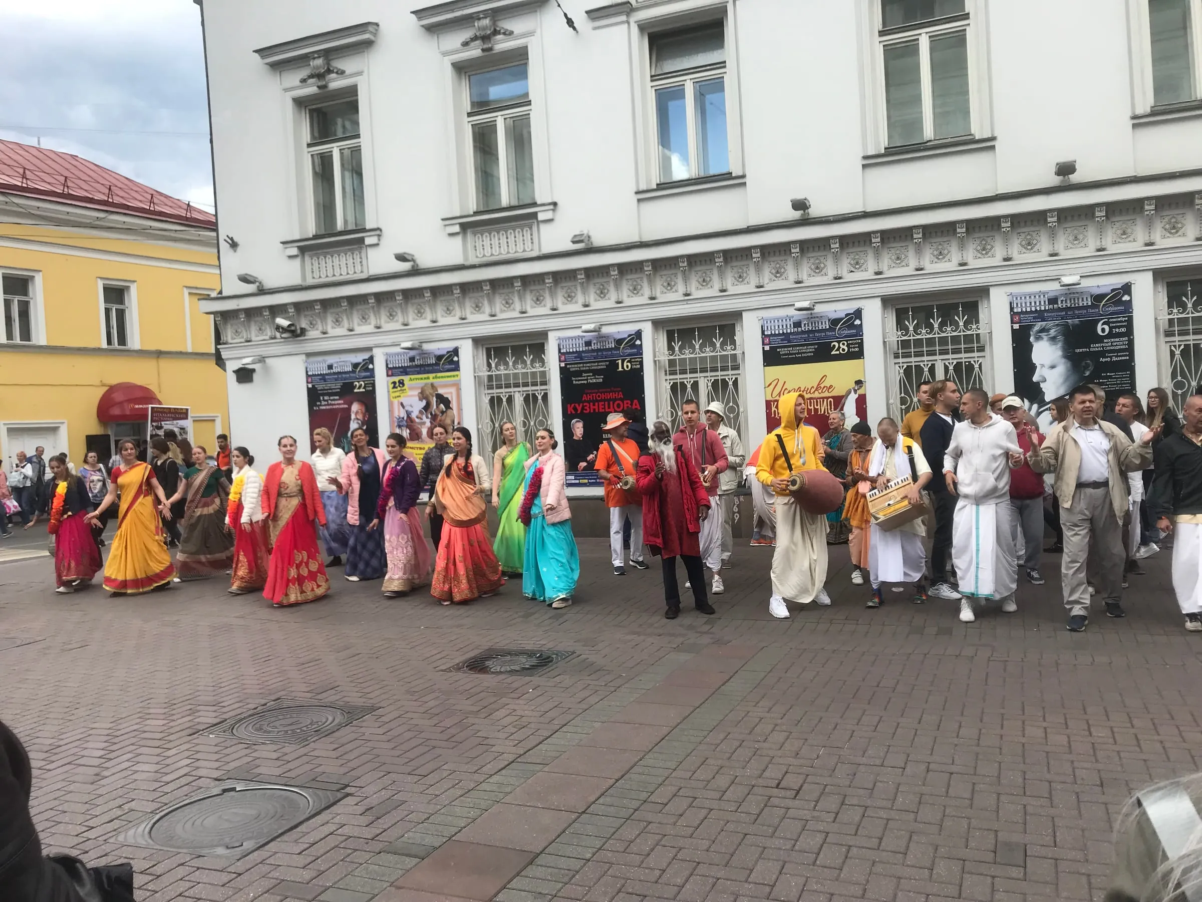 A colourful procession in saris and white clothes, singing and dancing along a pedestrian street