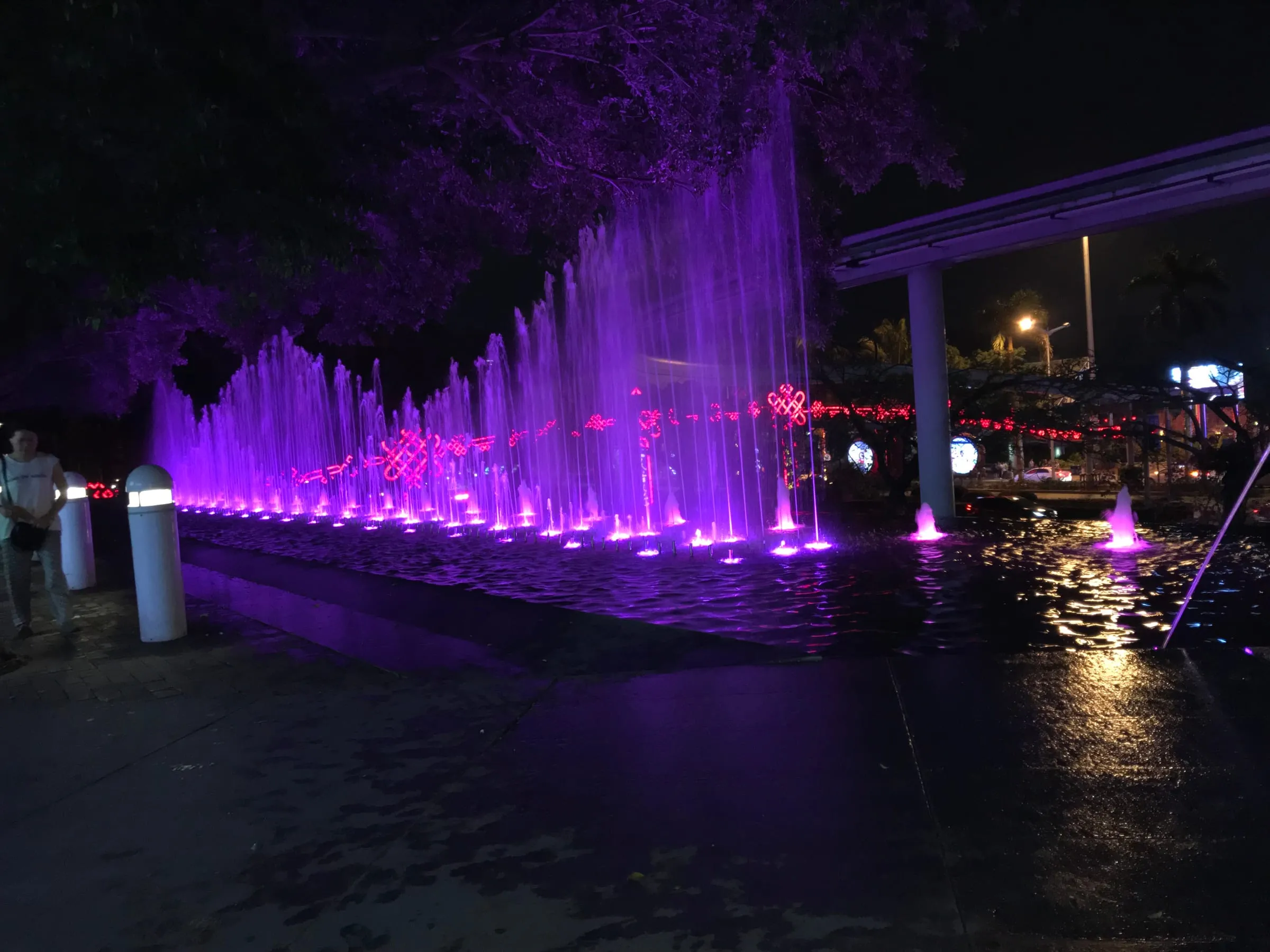 A wall of fountains lit purple at night, a monorail track crossing above