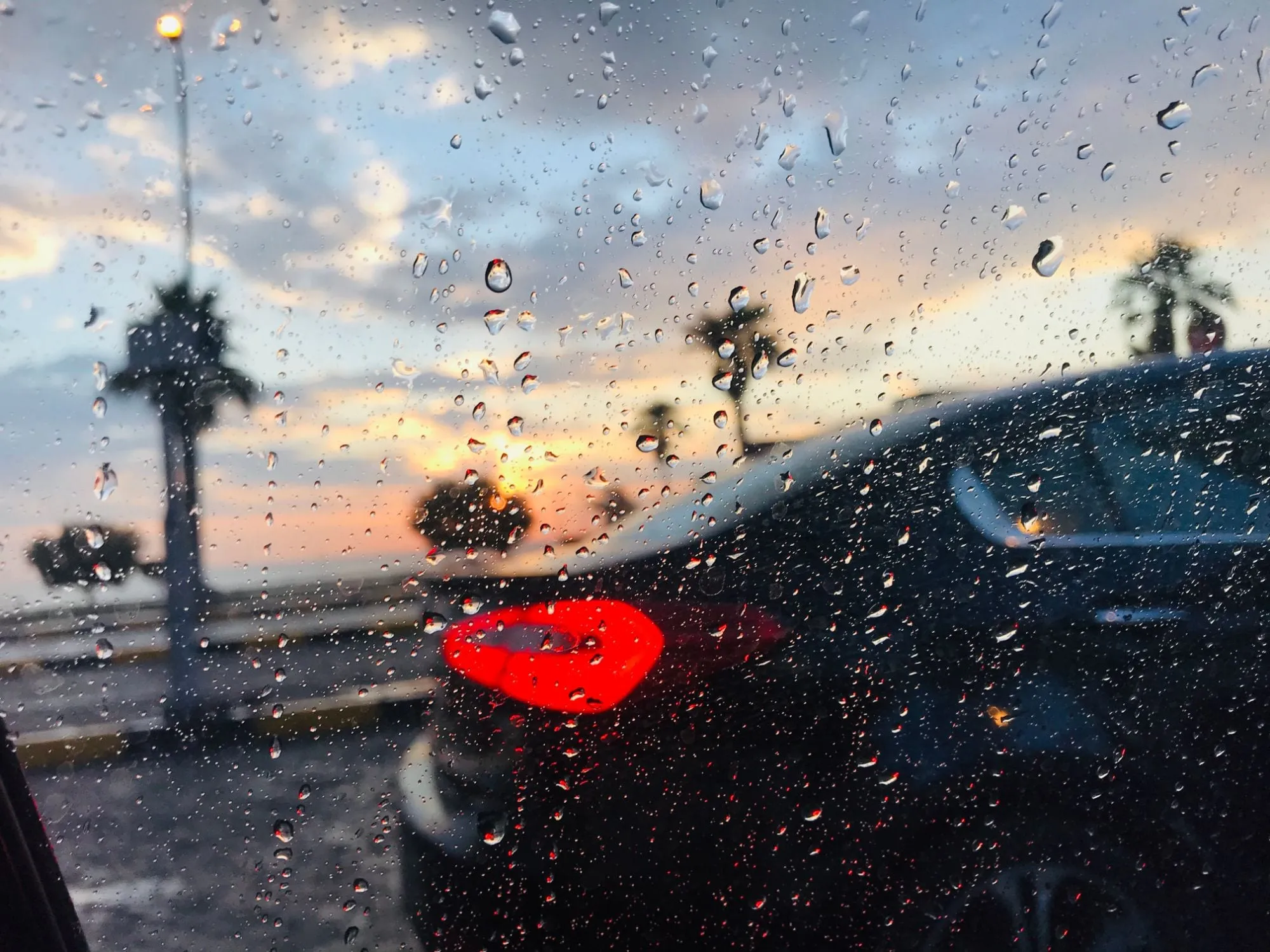 Rain drops on a car window at sunset, palm trees and a parked car blurred behind the glass, its red tail light glowing
