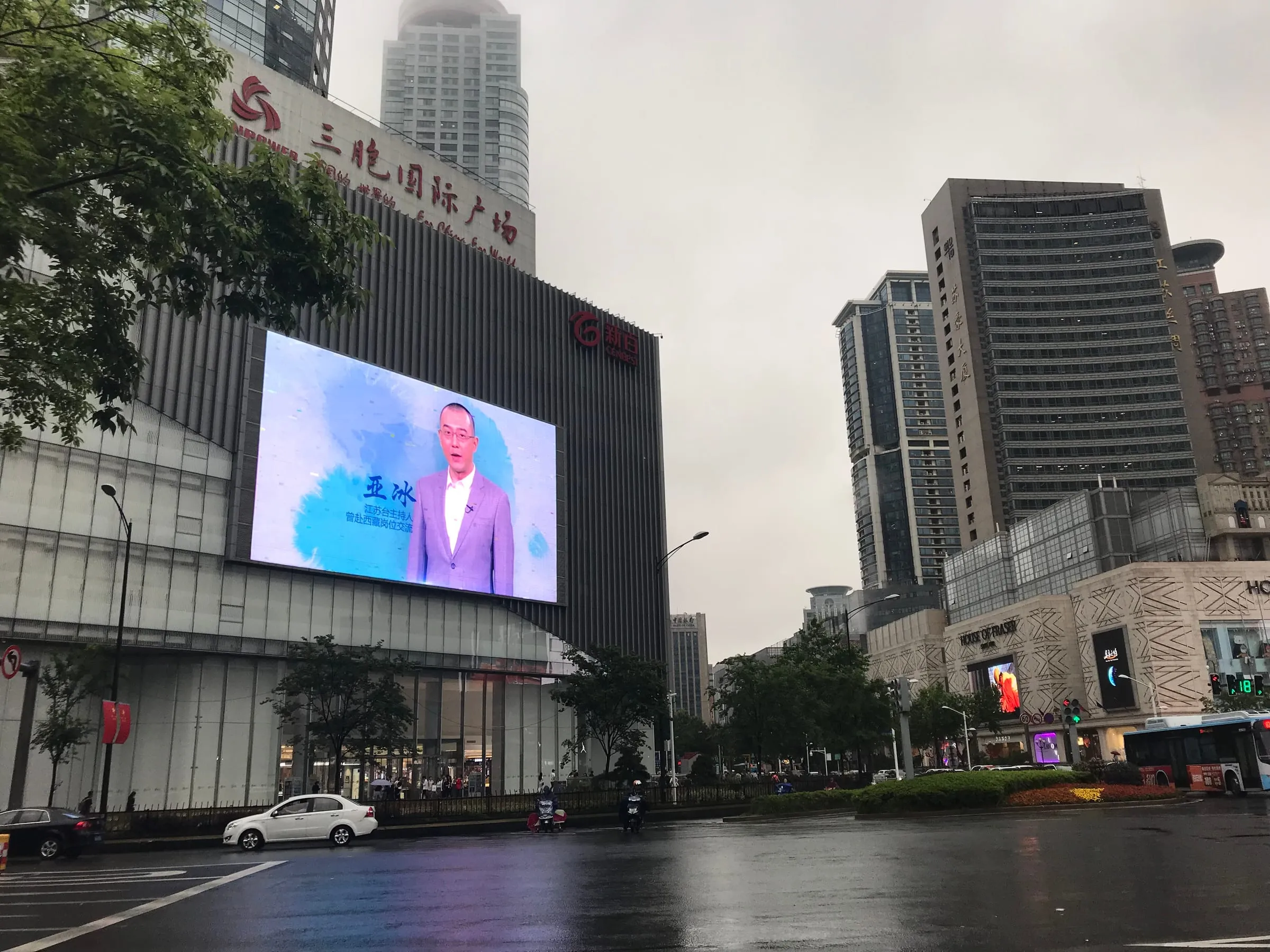 A wet street on a grey day, a large video screen on a shopping centre and towers beyond