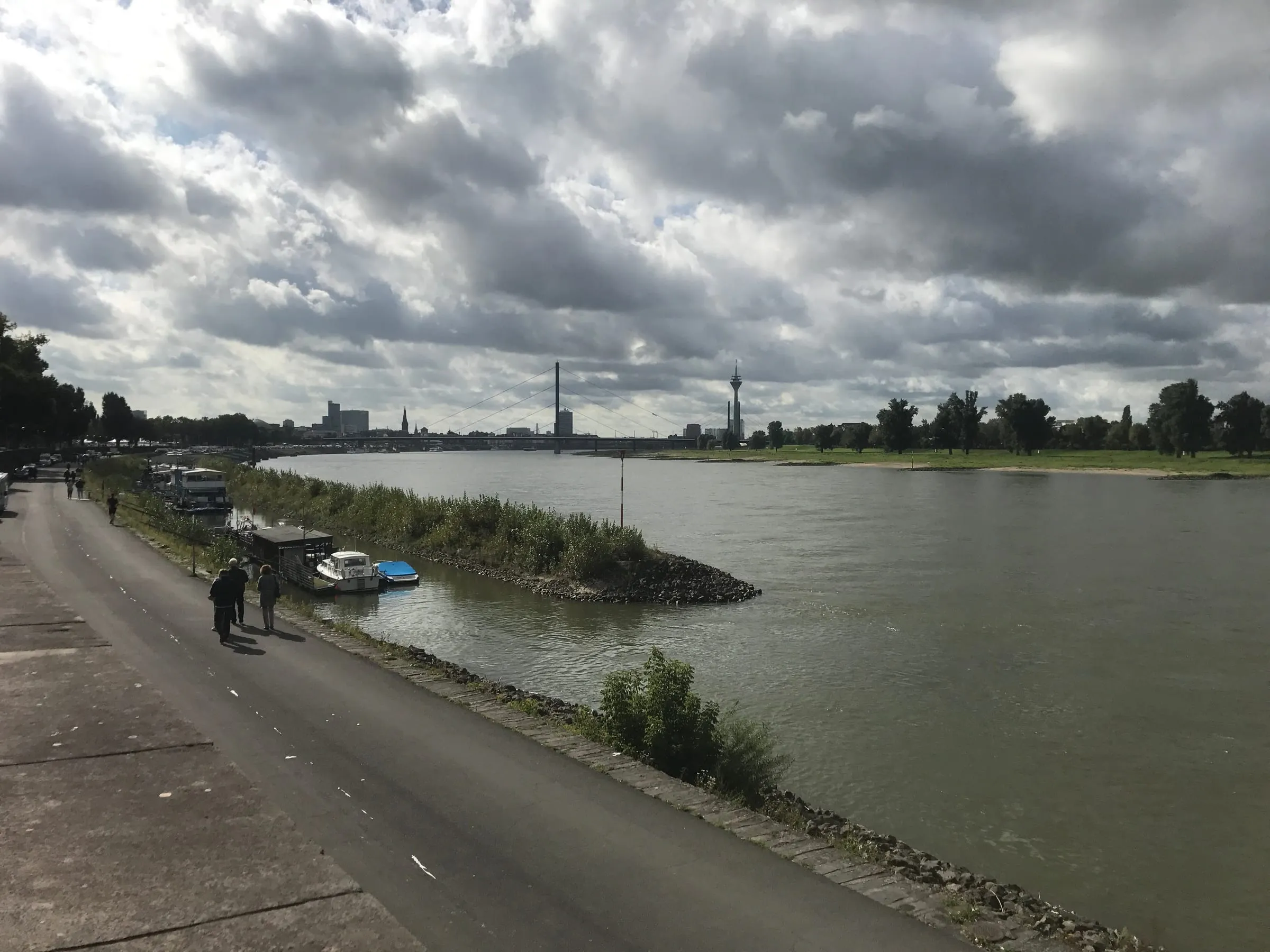 The wide Rhine under heavy clouds, a riverside path on the left and the city's tower far in the distance