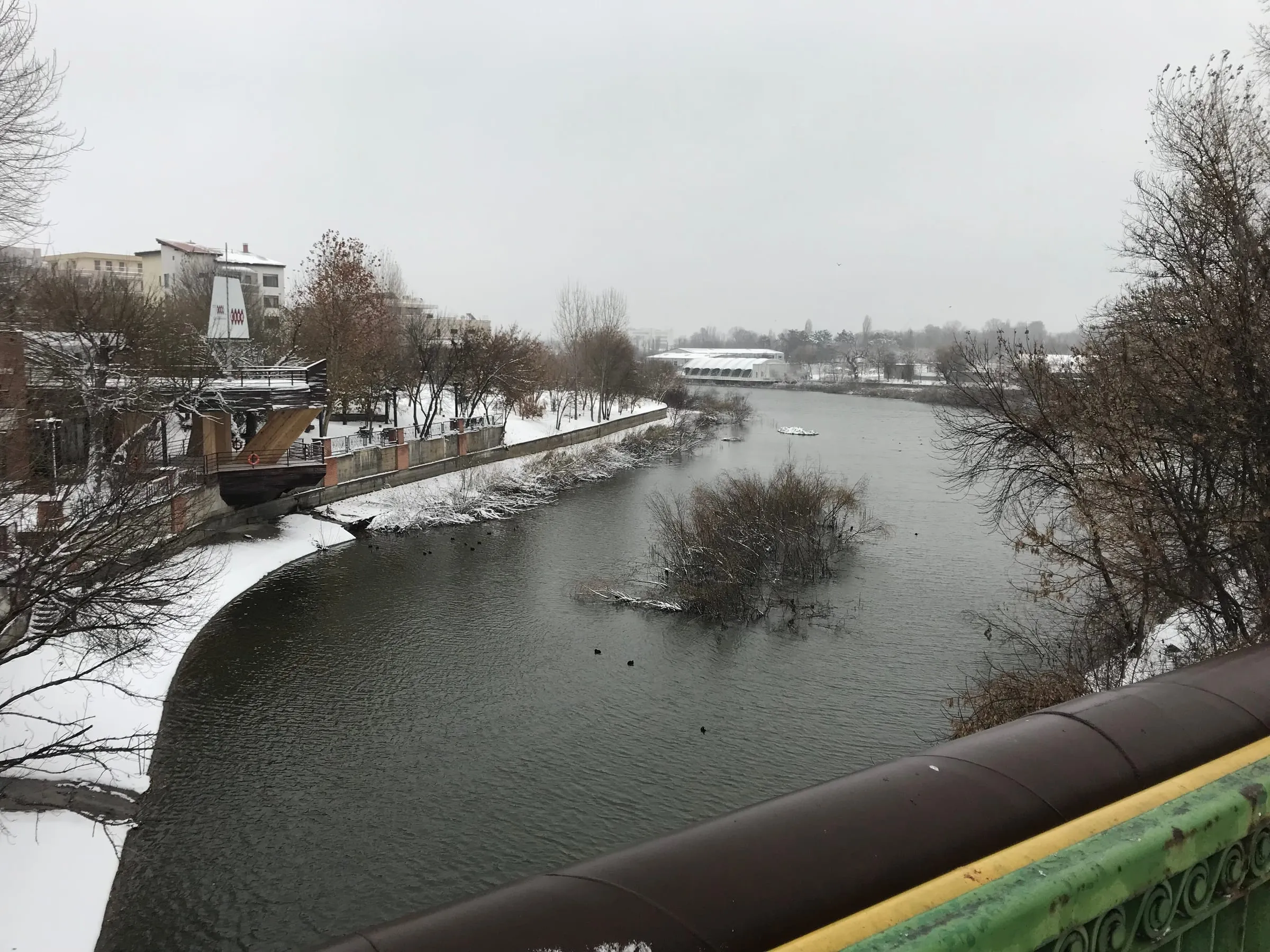 A grey river with snowy banks seen from a bridge, bare trees on both sides