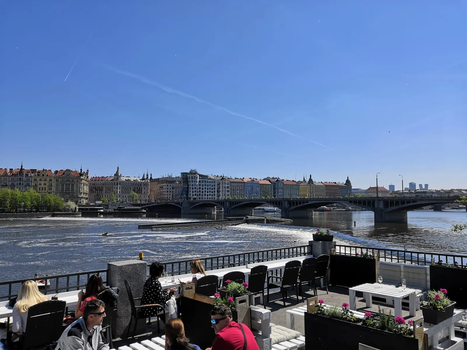 The river and a long bridge seen from a riverside terrace where people sit at tables on a clear day