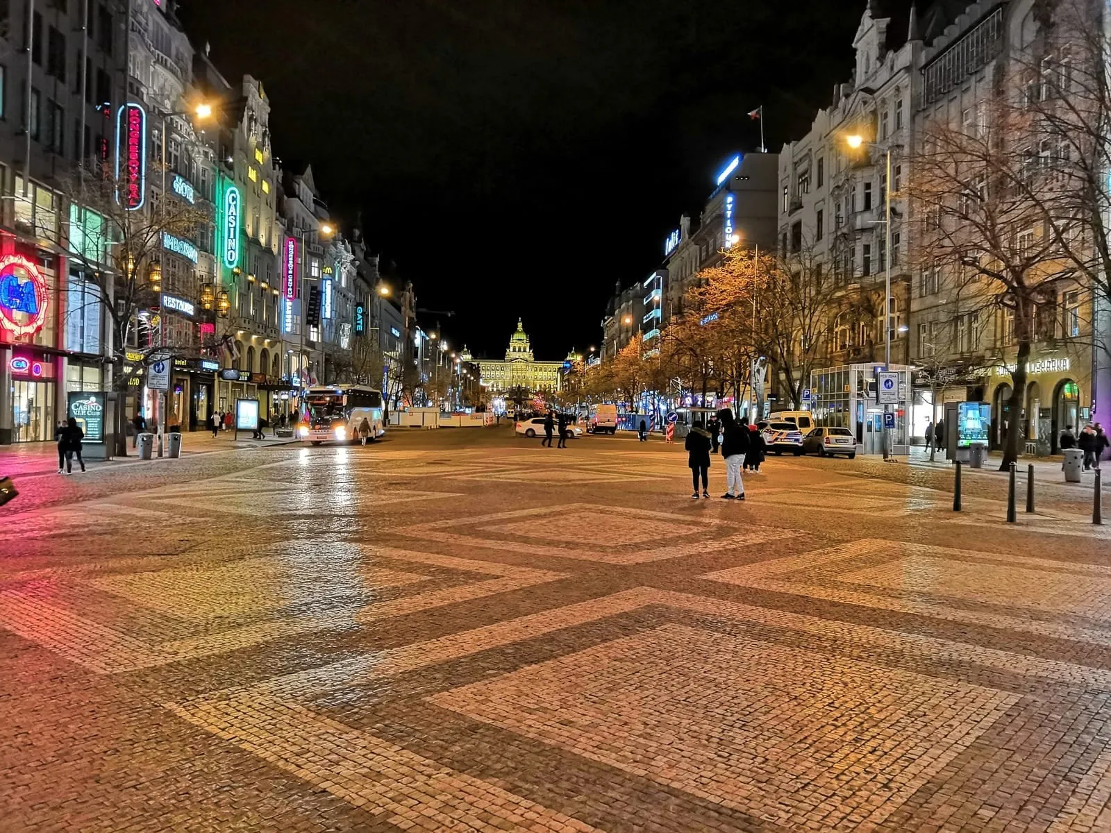 A long square at night with patterned paving, lit shops on both sides and a domed building at the far end