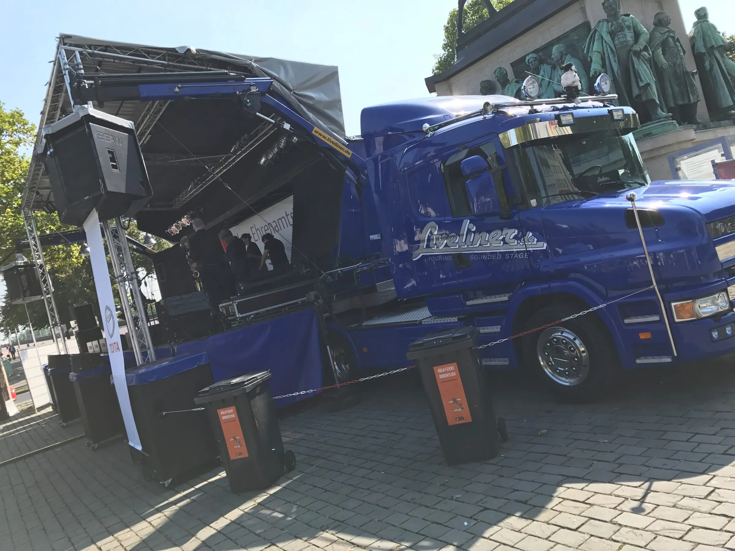 A blue truck opened up as a mobile stage on a cobbled square, a bronze statue behind it