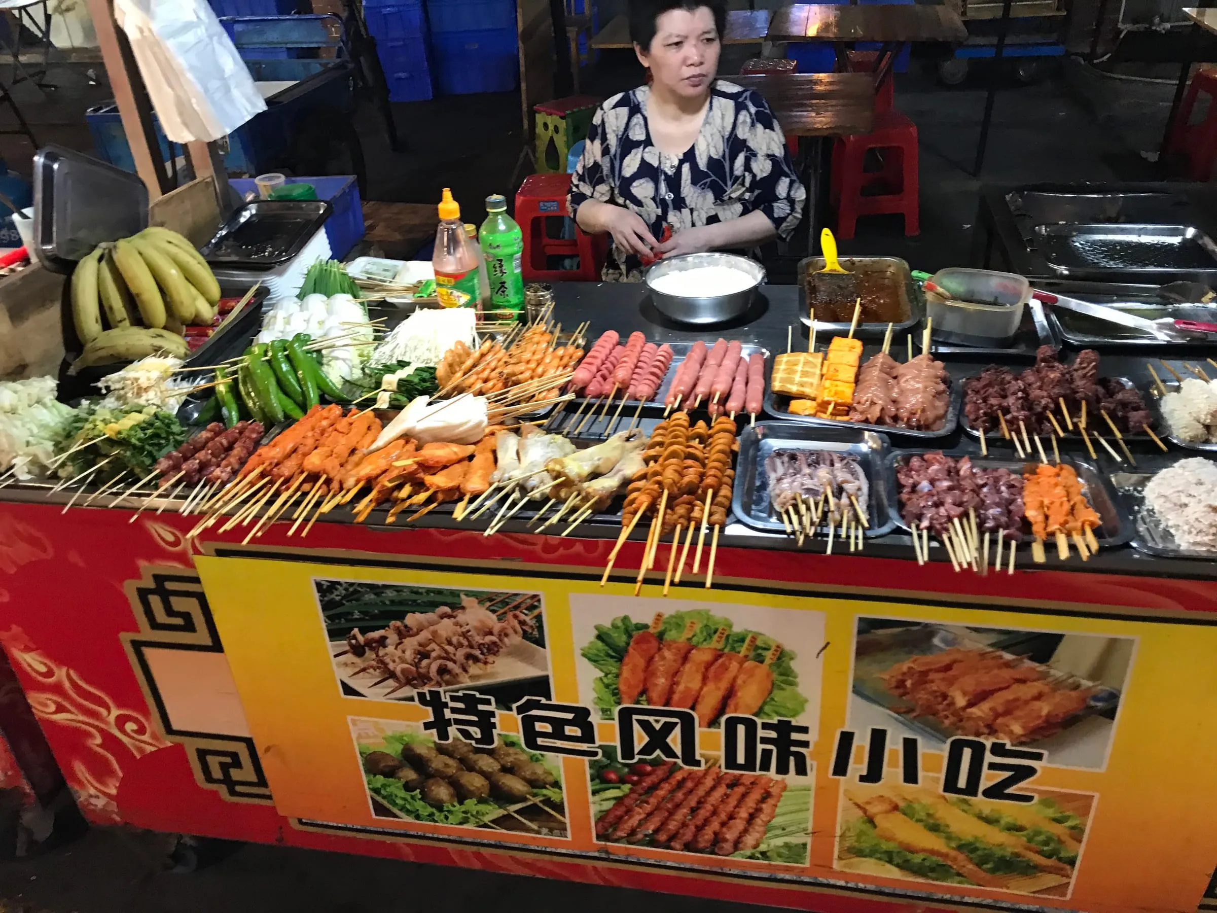A street food stall with skewers of meat, seafood and vegetables laid out, a woman waiting behind the counter
