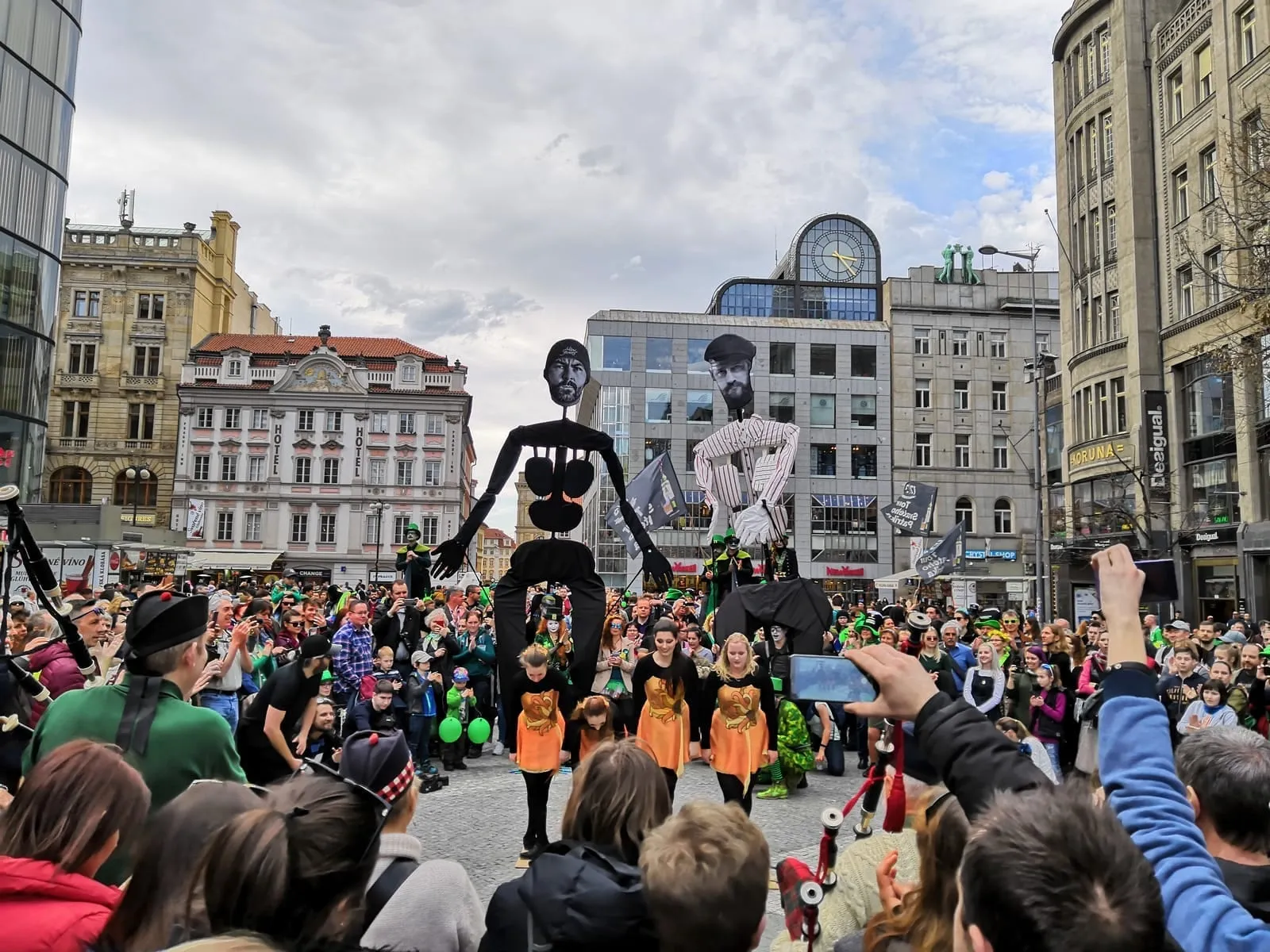 A crowd in a city square watching a street performance with tall puppet figures