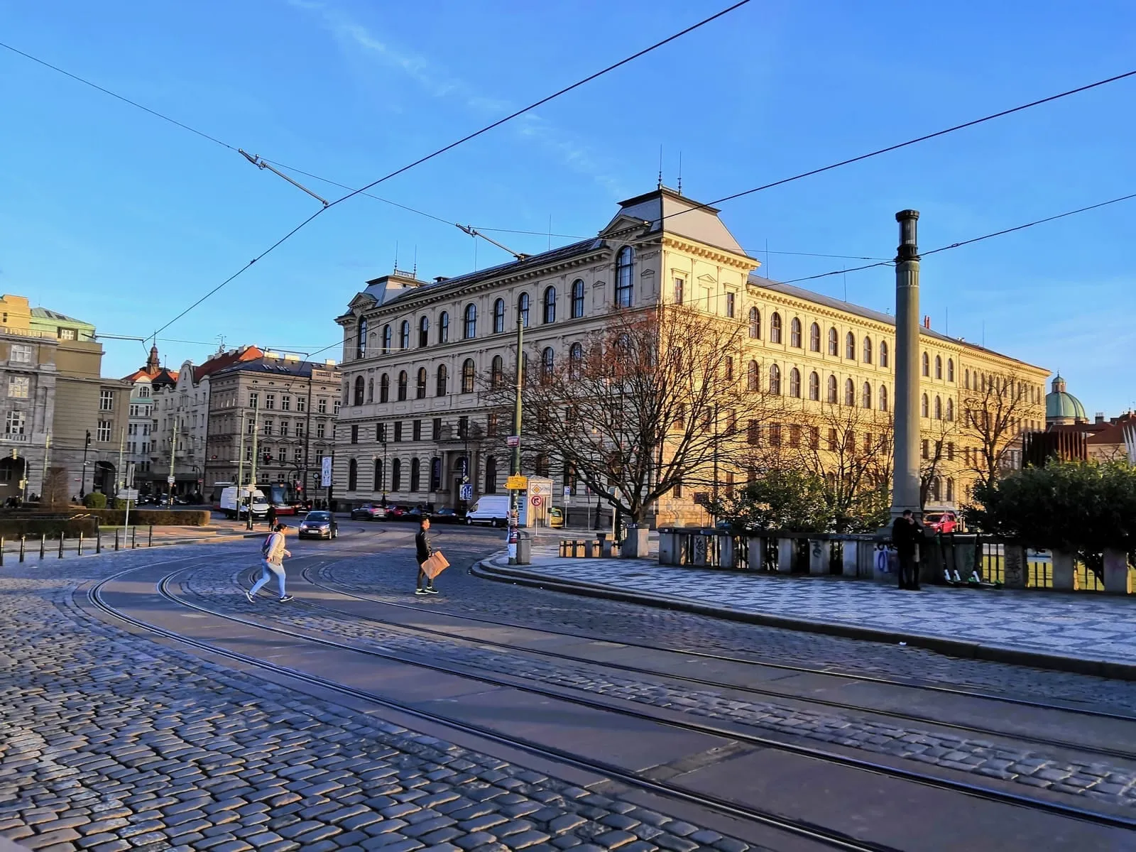 A wide cobbled street with tram lines, a large stone building and a tall column under a blue sky