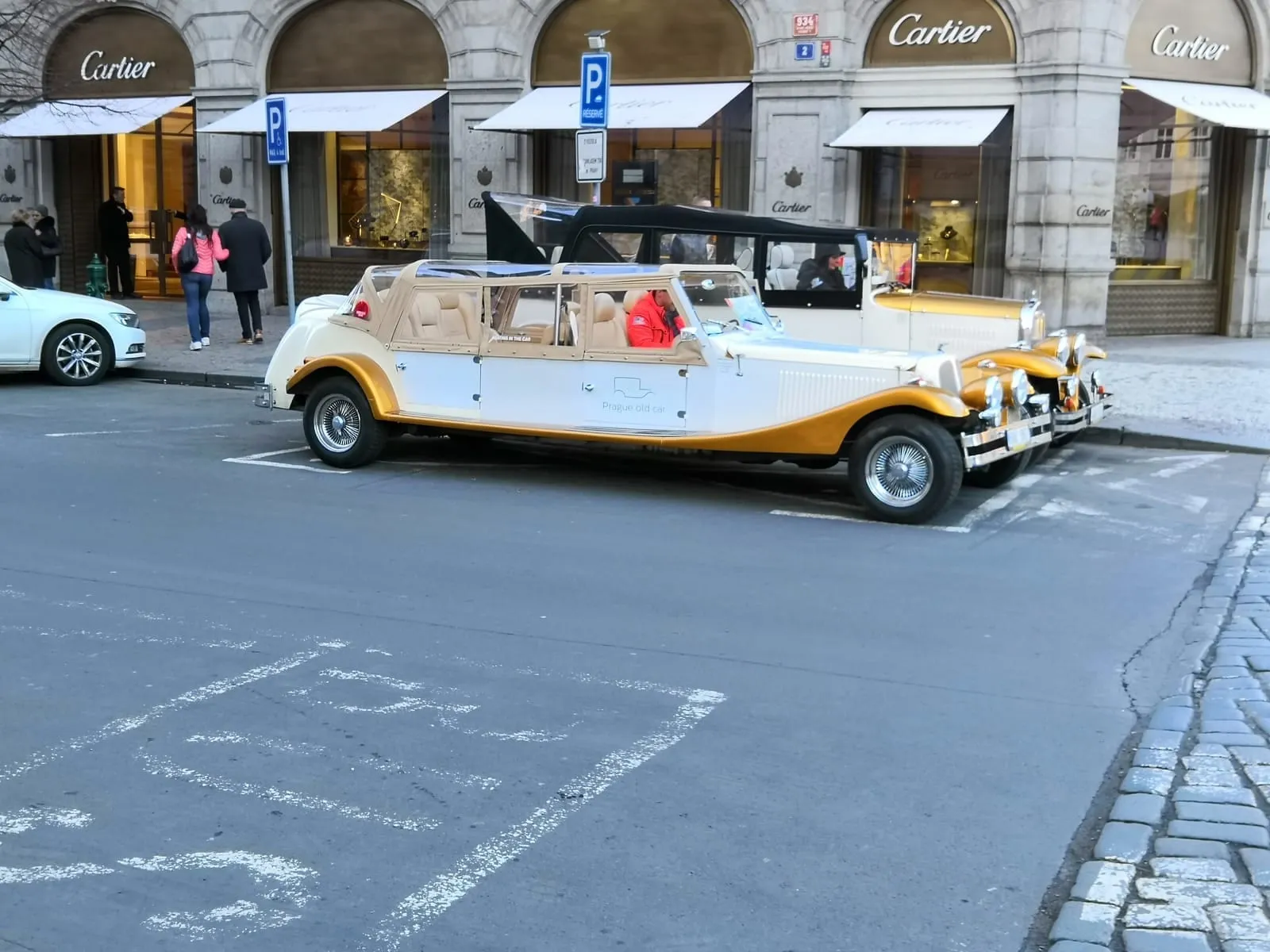 A cream and gold vintage-style open car parked on a cobbled street in front of shop fronts