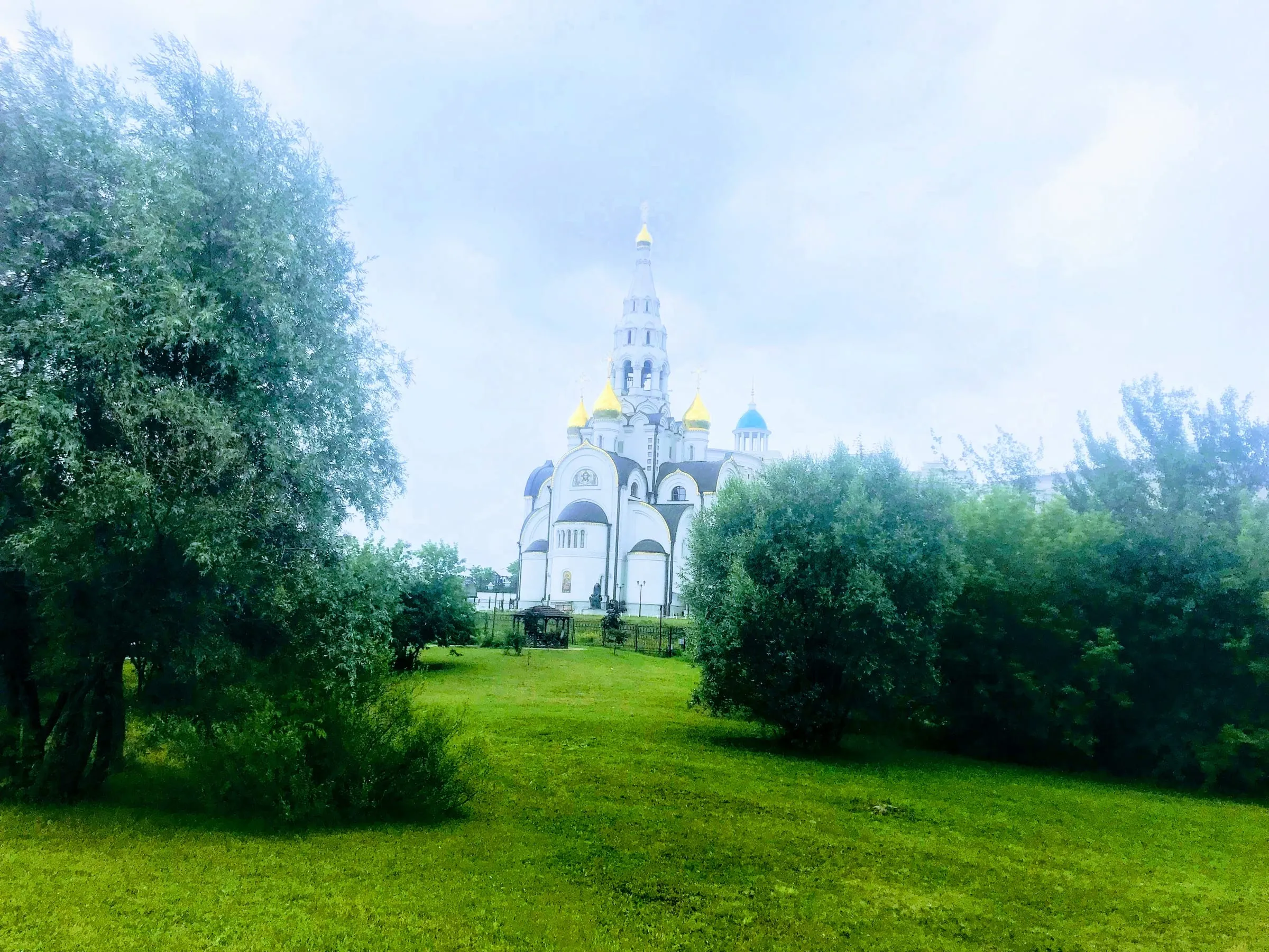 A white church with golden domes beyond a green lawn and full trees